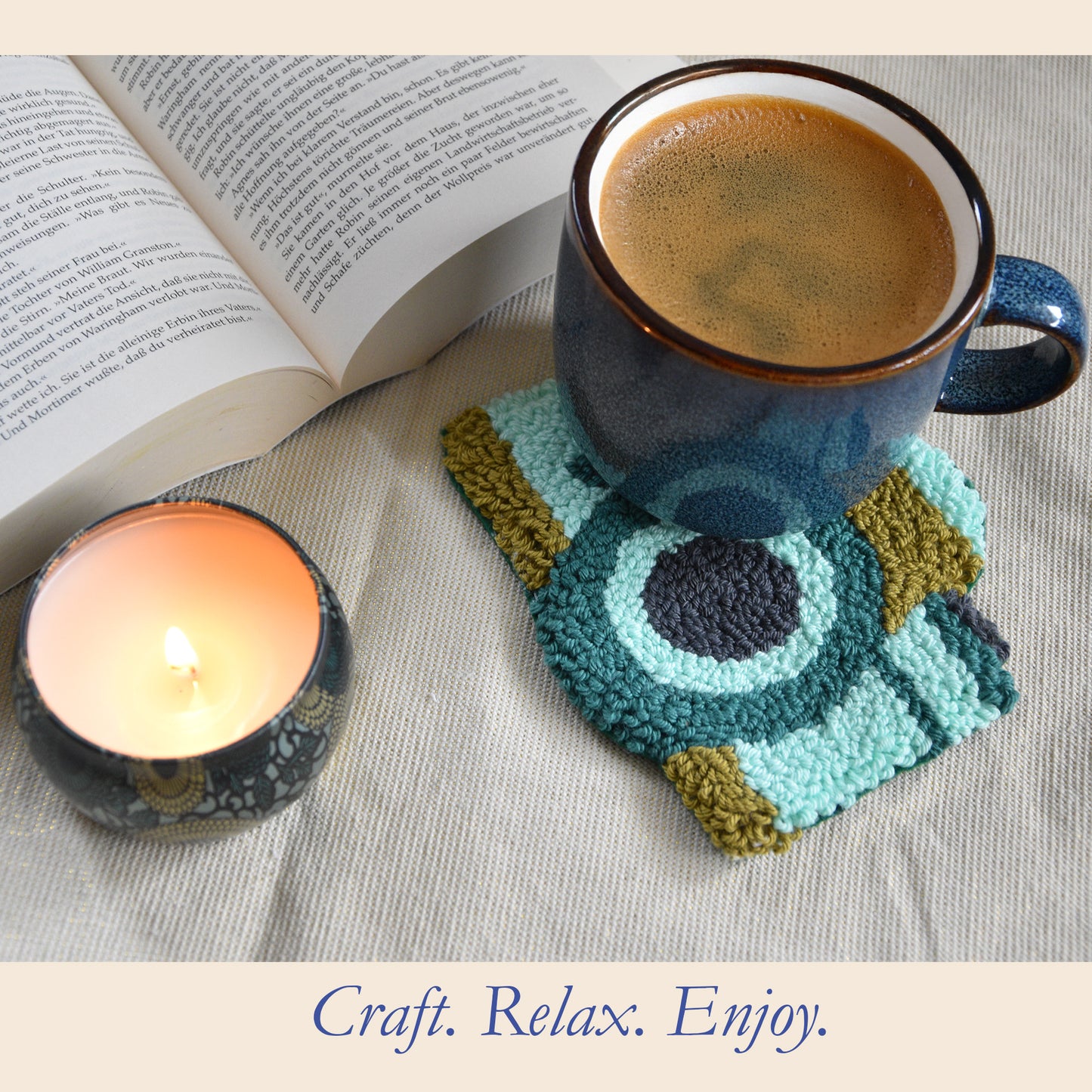 A cozy lifestyle scene featuring a punch needled coaster with a retro camera in teal, light blue, and olive green, holding a dark blue ceramic mug filled with coffee. An open book and a lit candle are placed nearby on a textured beige fabric background, with the text 'Craft.