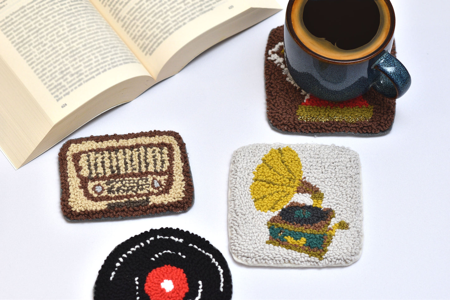 Set of handmade punch needle coasters with vintage designs on a white surface with an open book and mug.
