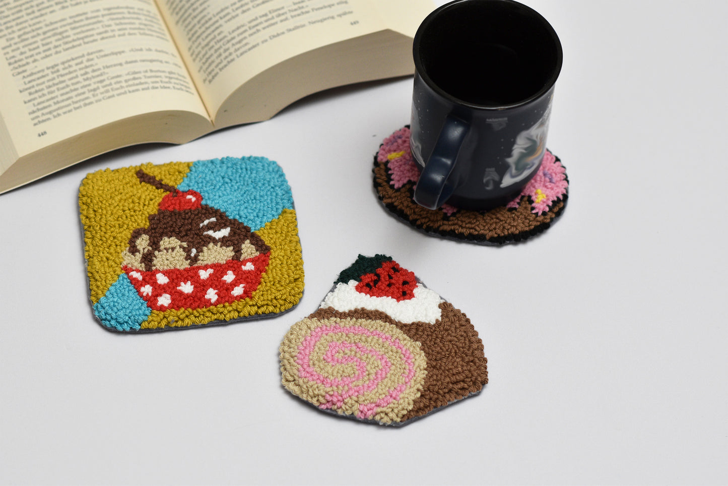 Three colorful coasters with dessert designs on a white surface next to an open book and a black mug.