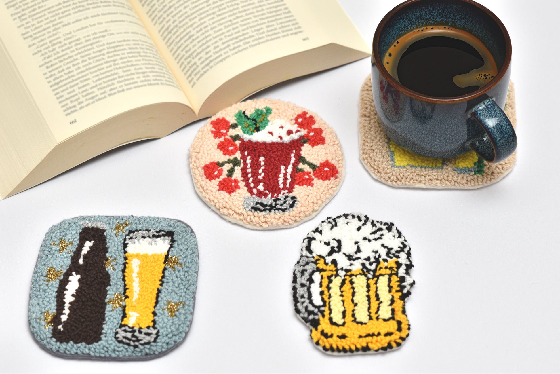 Set of coasters with designs of a beer bottle, glass, and cherry drink on a white surface with a book and mug.
