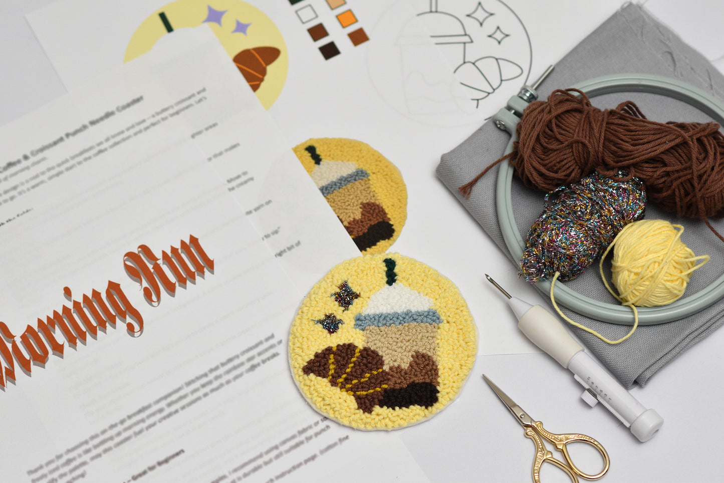 Embroidery hoop and a completed punch needle coaster with coffee and croissant design, yarn, scissors, and pattern sheet on a white surface.