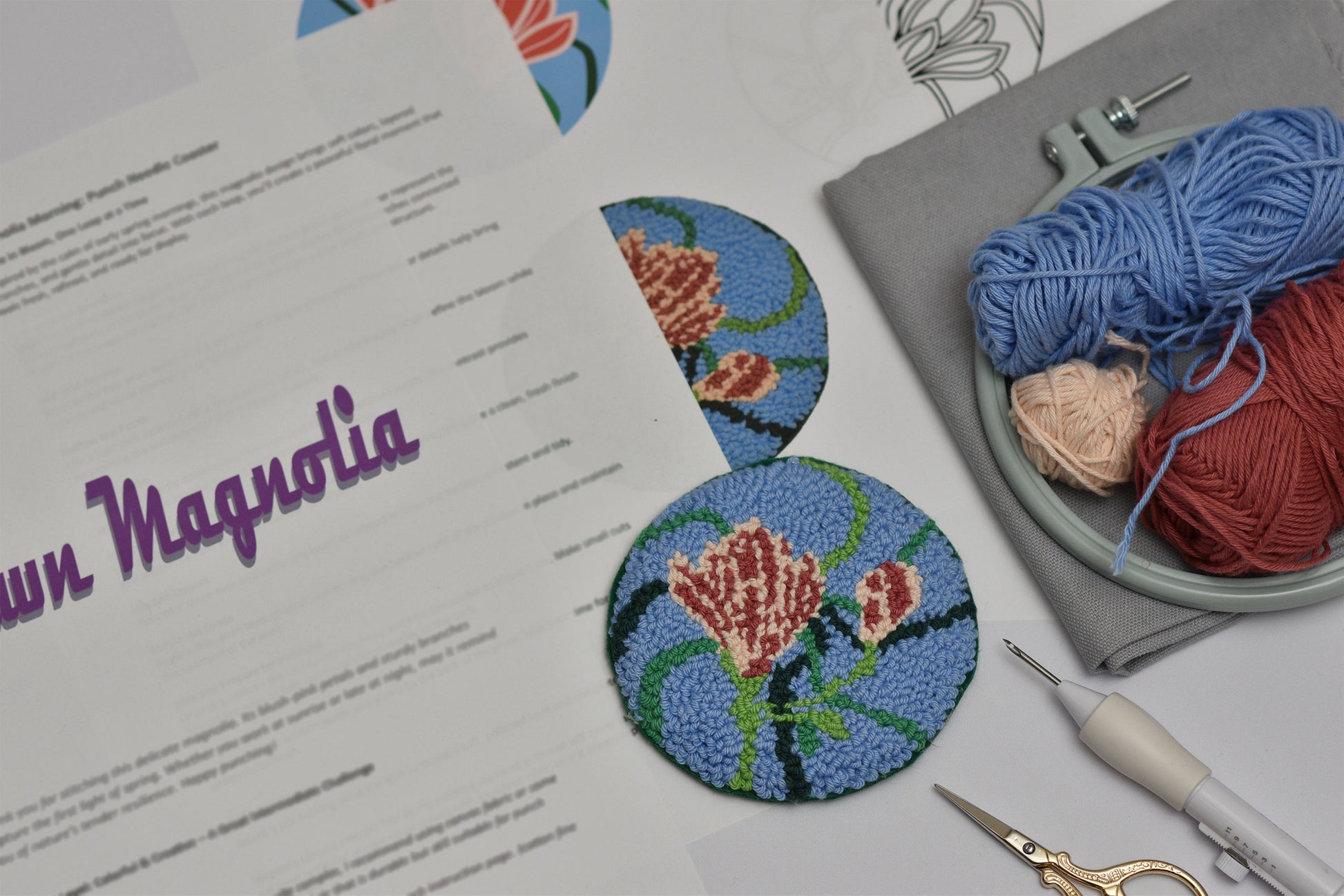 Embroidery hoop with floral magnolia design, yarn, scissors, and pattern sheet on a flat surface.