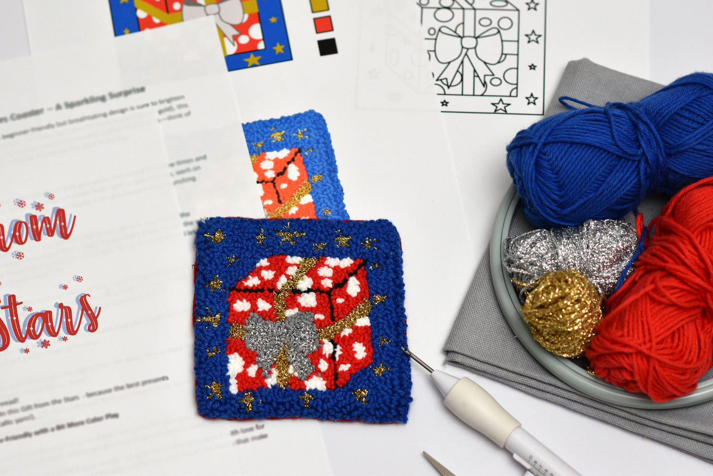 Decorative punch needle coaster with a red and gold design gift package on a blue background, surrounded by yarn and crafting tools.
