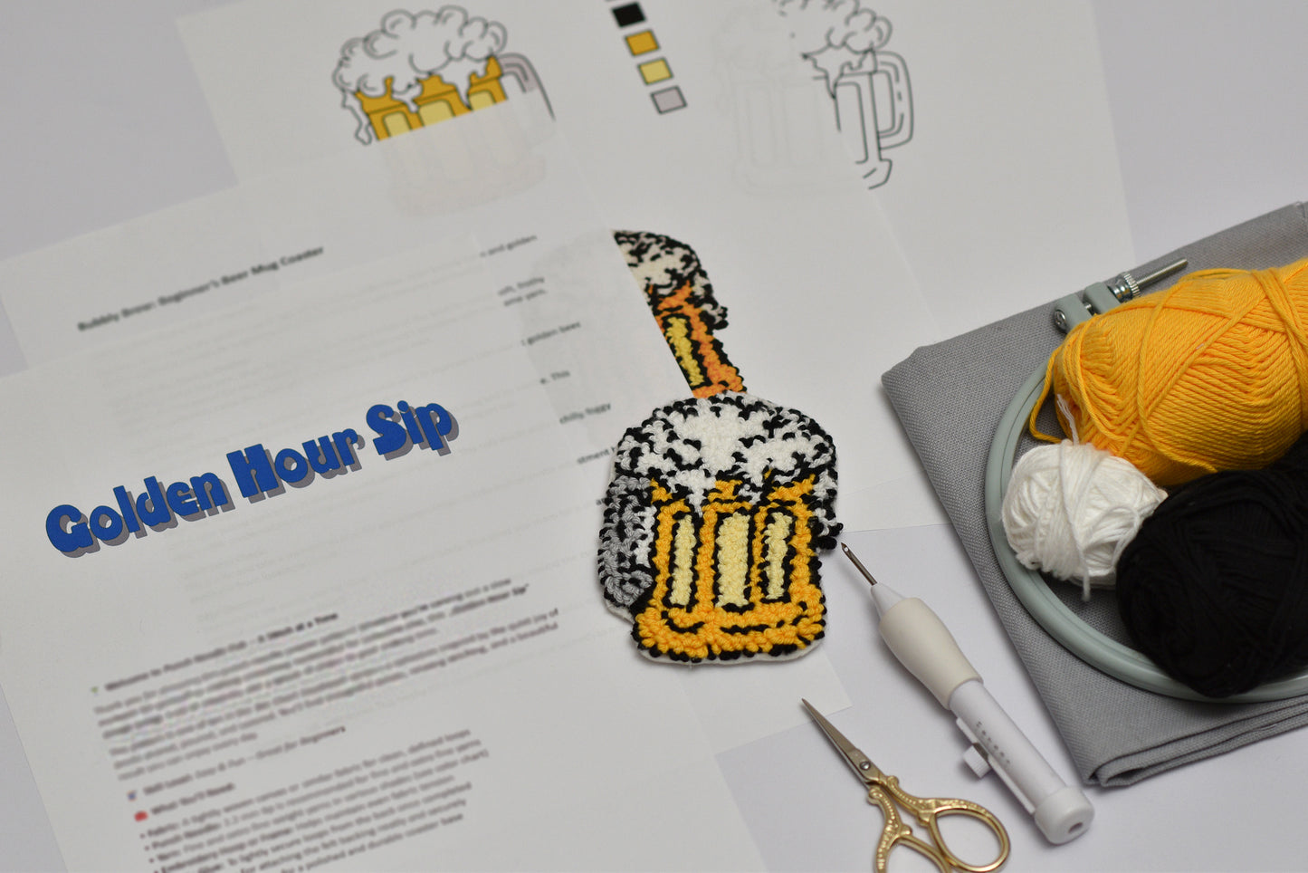 Punch needled beer mug with foam on a piece of paper, surrounded by yarn and embroidery tools.