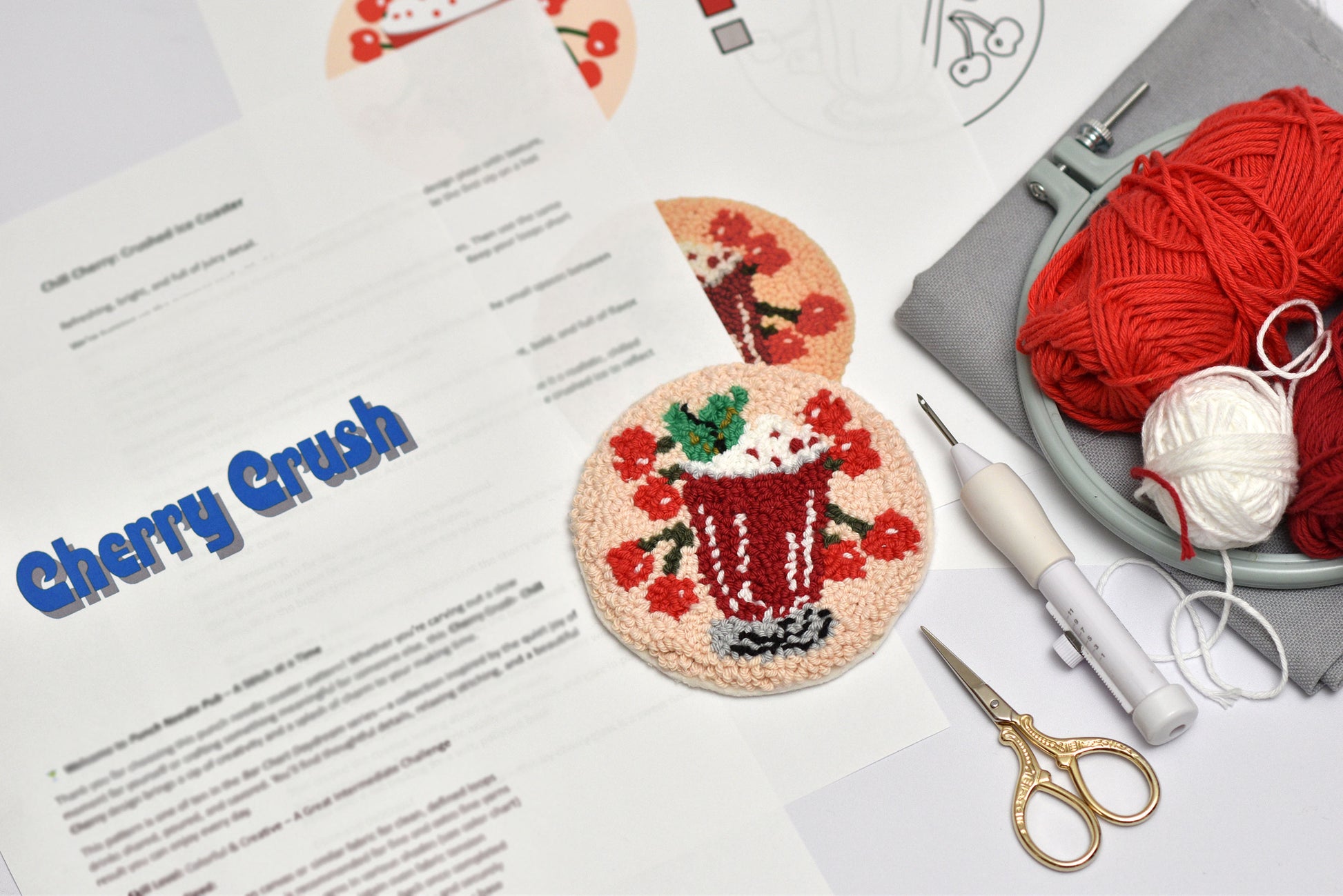 Punch needle coaster with cherry-themed design on a table with yarn, scissors, and pattern sheet.
