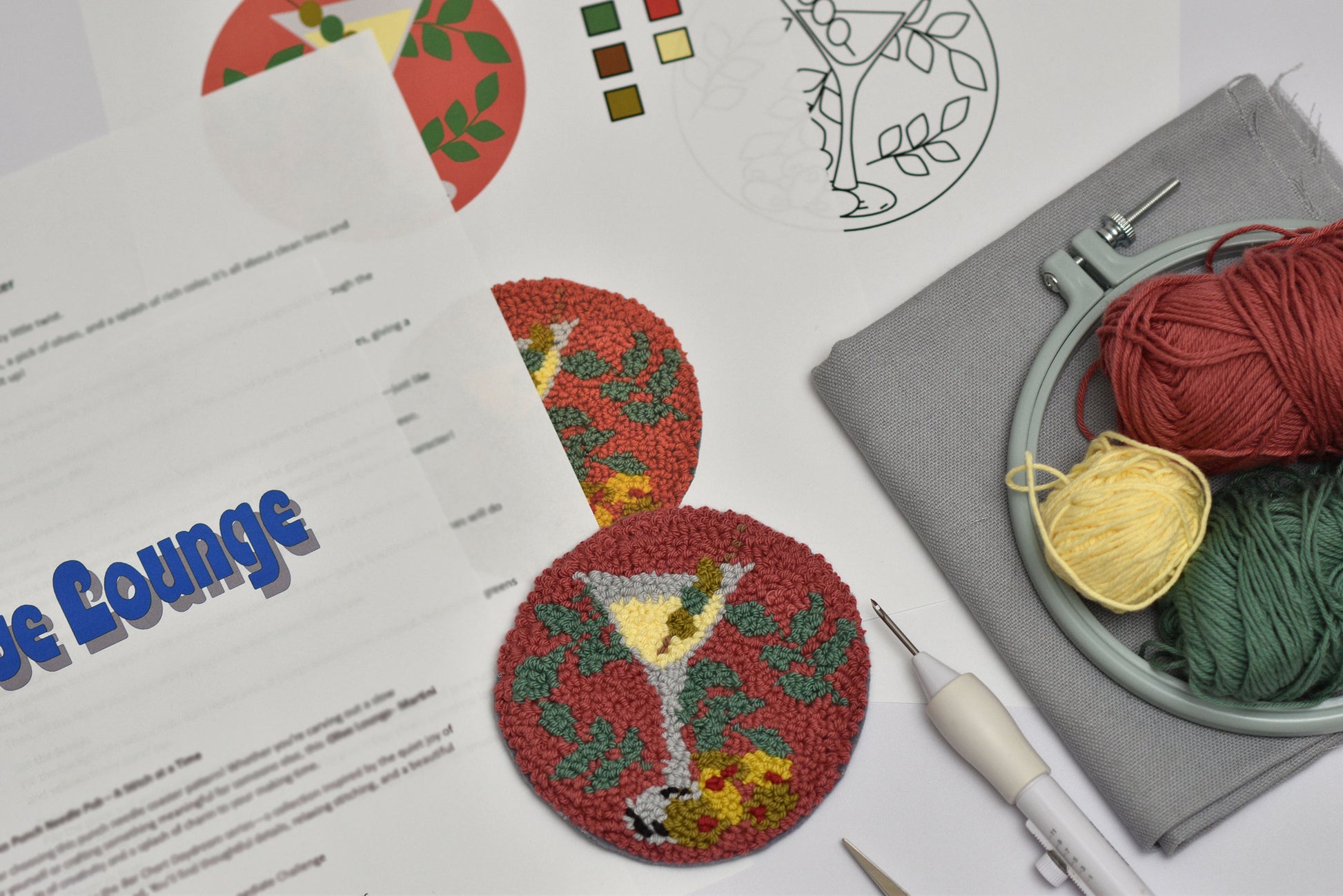 Embroidery hoop, embroidered coaster with Martini design, yarn, embroidery pattern, and stitching tool on a table.