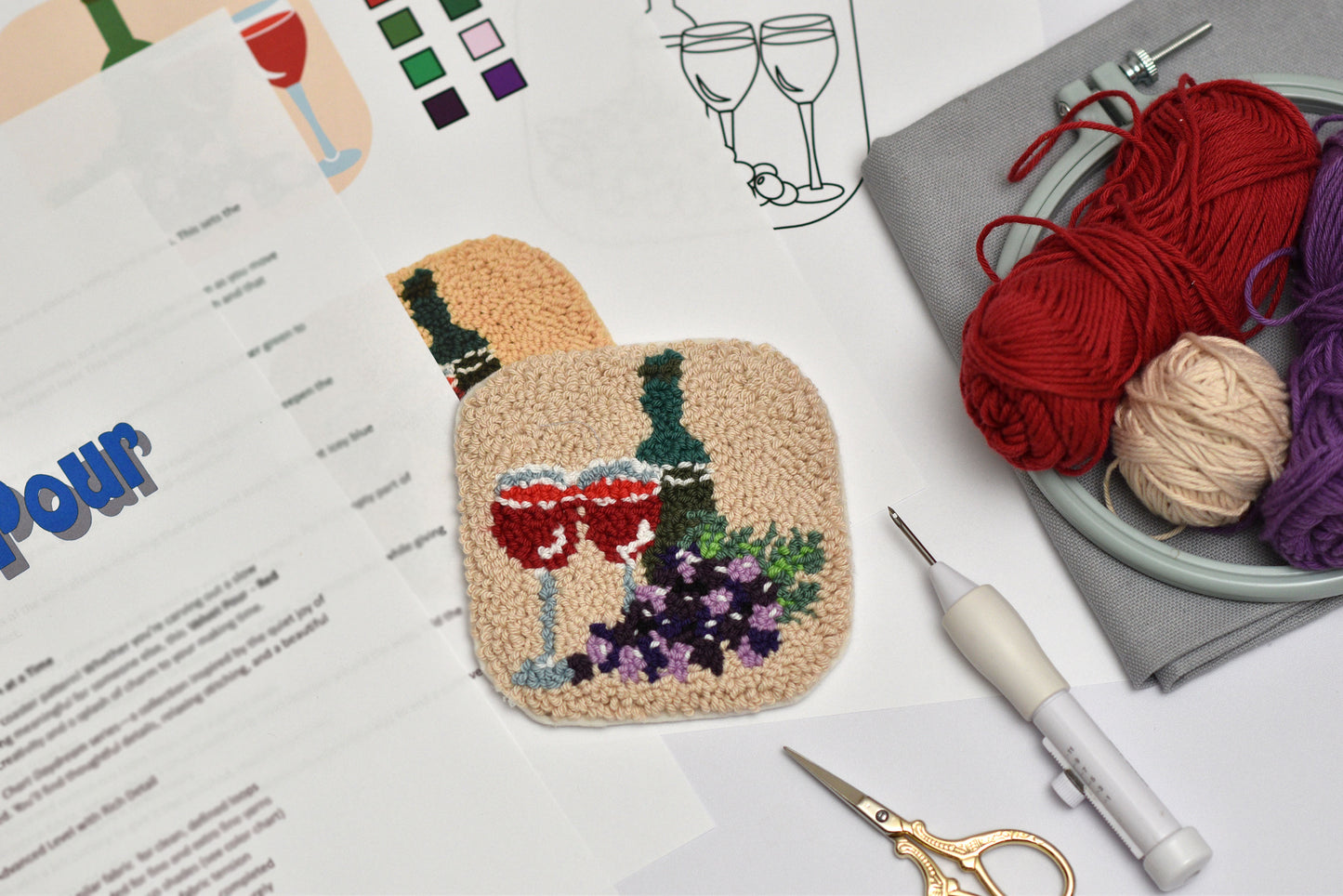 Embroidered coaster of a wine bottle, glasses and grapes on a white surface with yarn and tools.