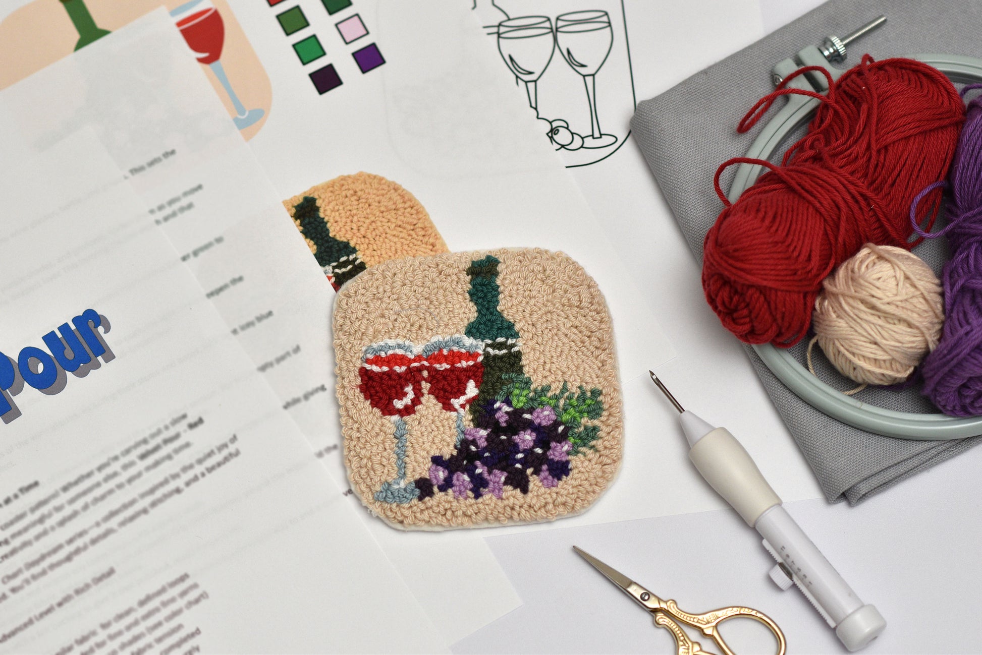 Embroidered coaster of a wine bottle, glasses and grapes on a white surface with yarn and tools.