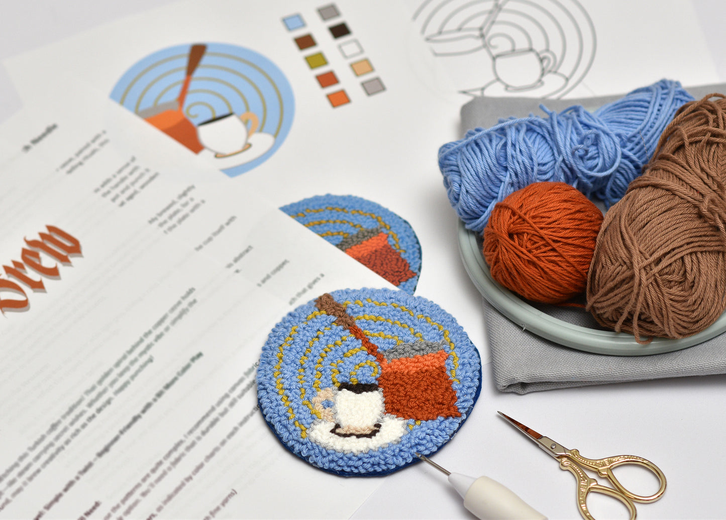 Embroidered coaster of a Turkish coffee pot with yarn balls on a table with yarn and scissors.