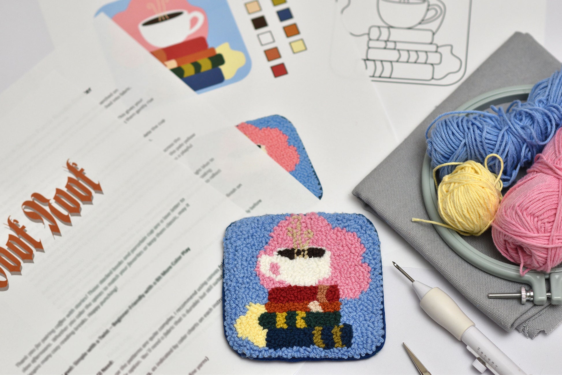 Embroidery project with a completed coaster with design of books and coffee cup, yarn, and embroidery tools on a table.