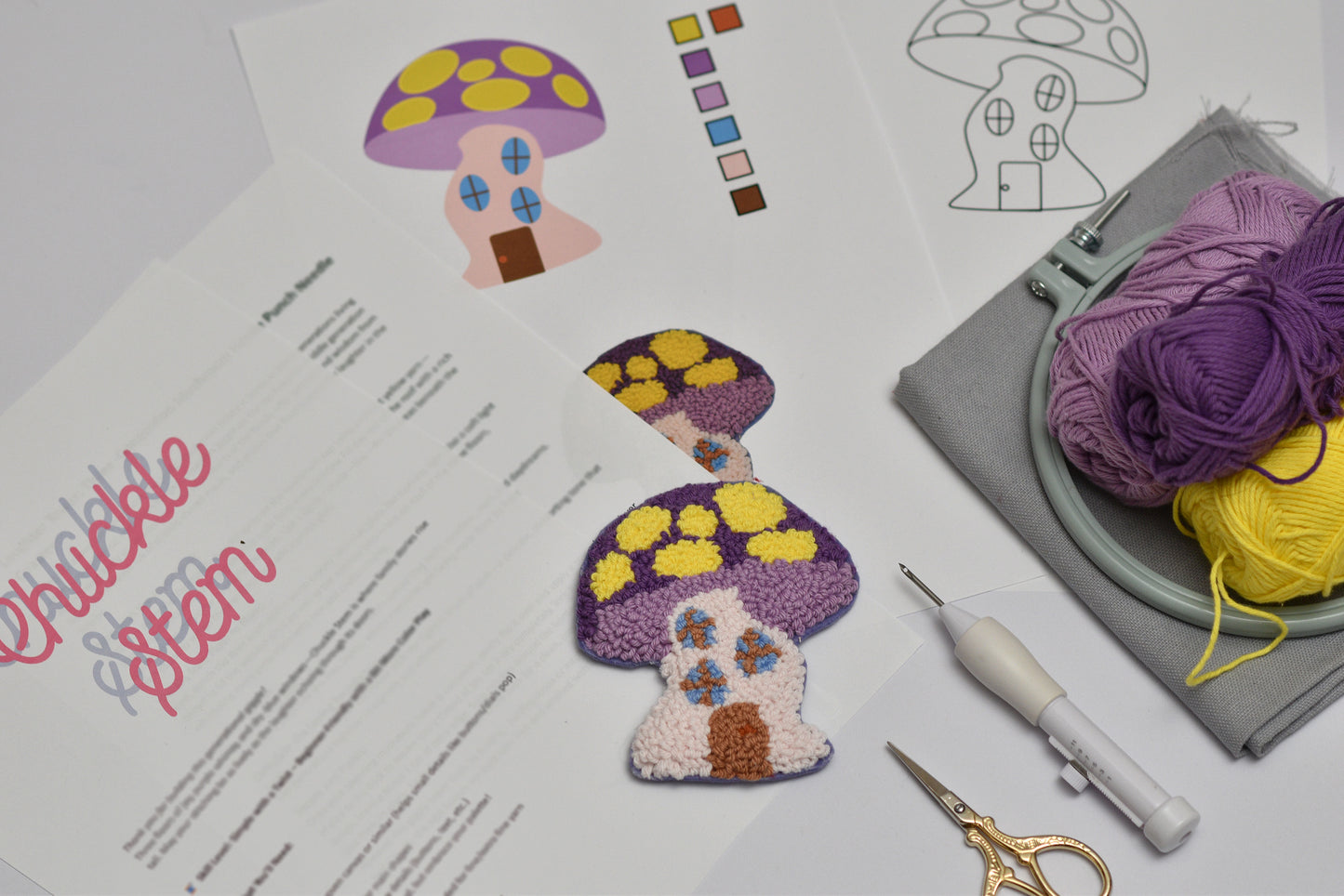 Embroidery coaster project with mushroom house design, yarn, and sewing tools on a table.