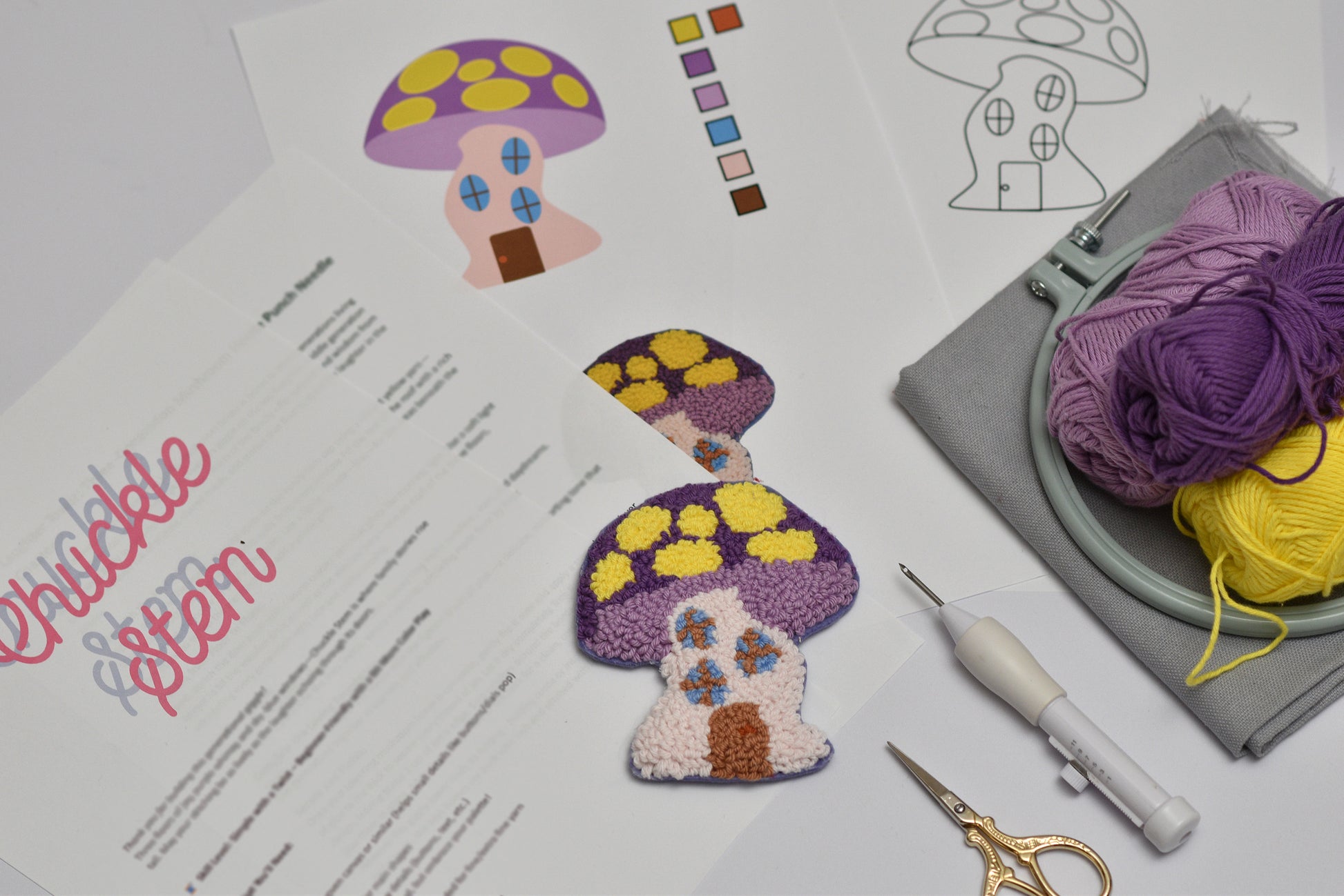 Embroidery coaster project with mushroom house design, yarn, and sewing tools on a table.