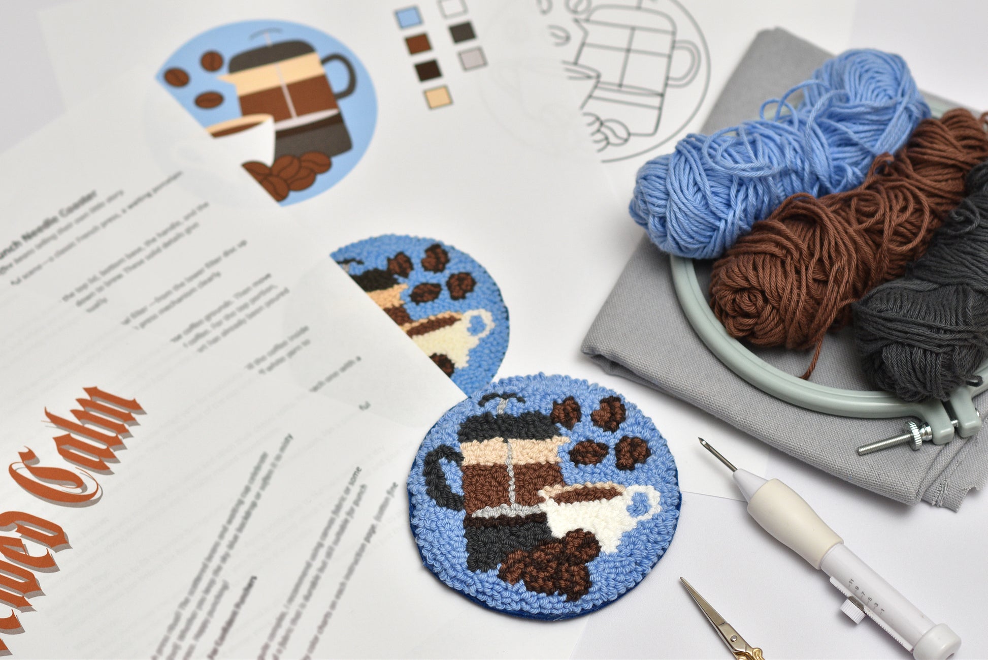 Punch needled coaster with French press coffee-themed design, yarn, and tools on a table.