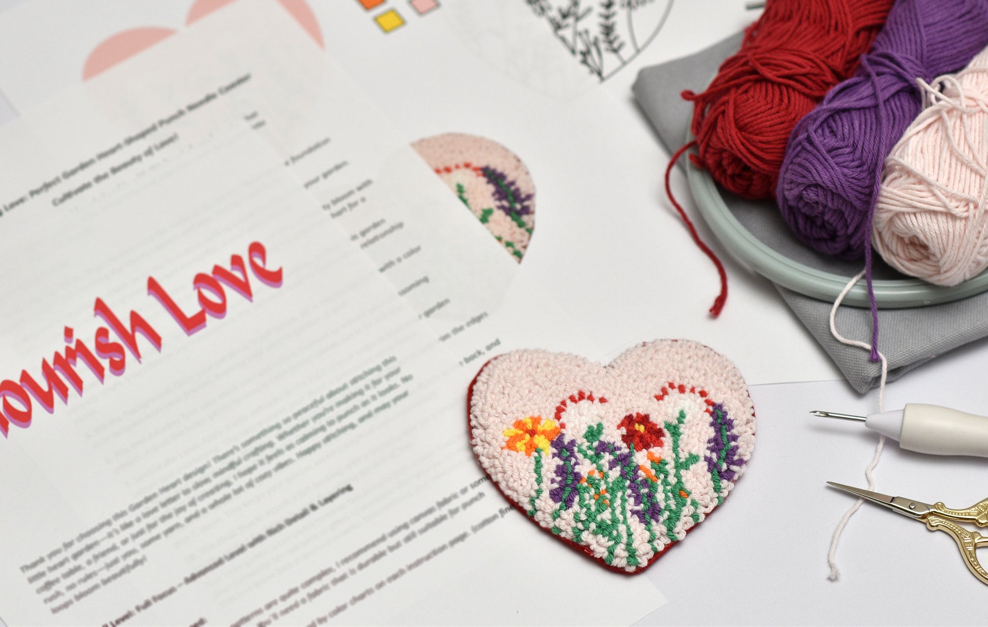 Embroidered heart with floral design on a printed sheet of text, surrounded by yarn and sewing tools.