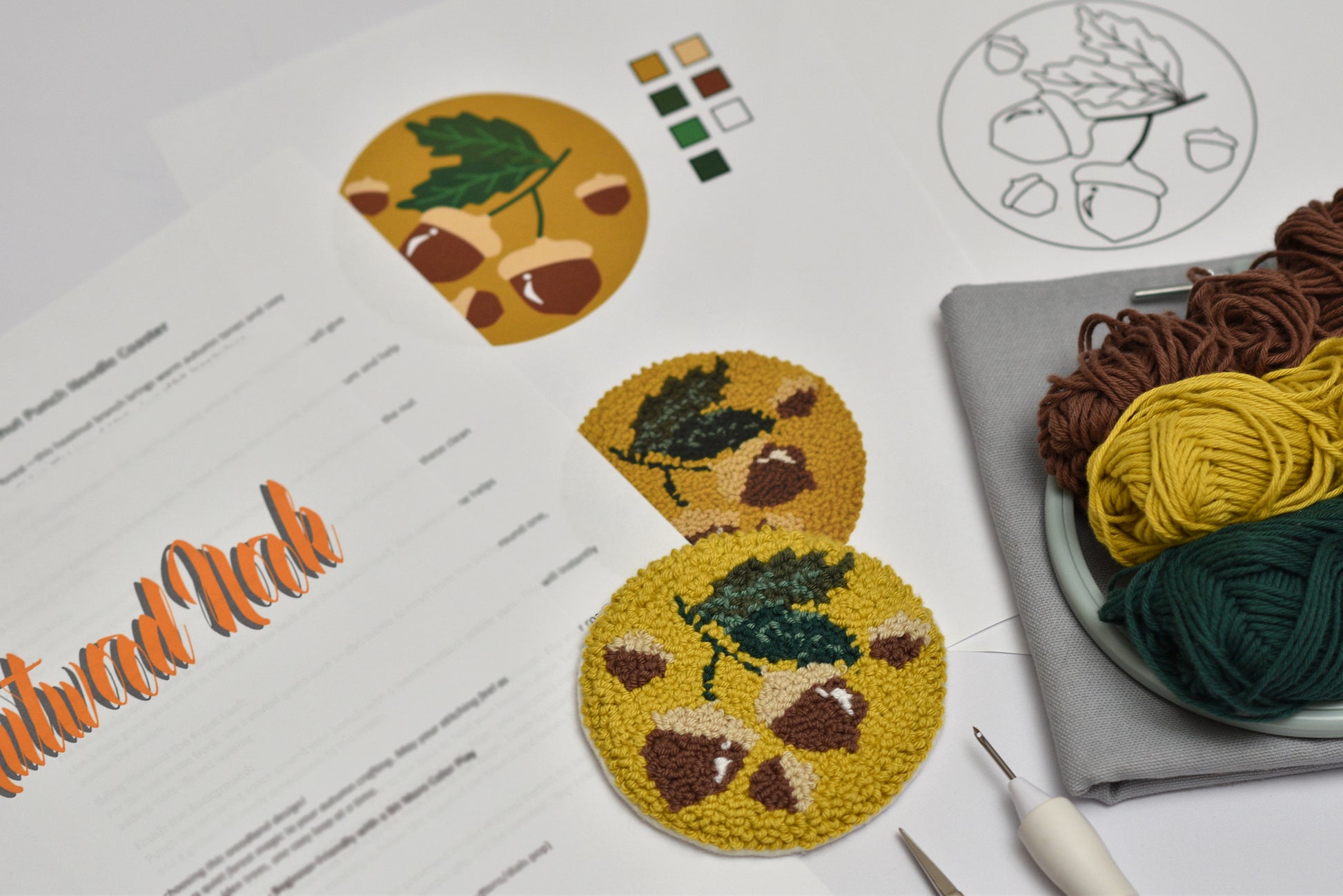 Embroidery coaster project with a hazelnut and leaf design, yarn balls, and embroidery tools on a table.