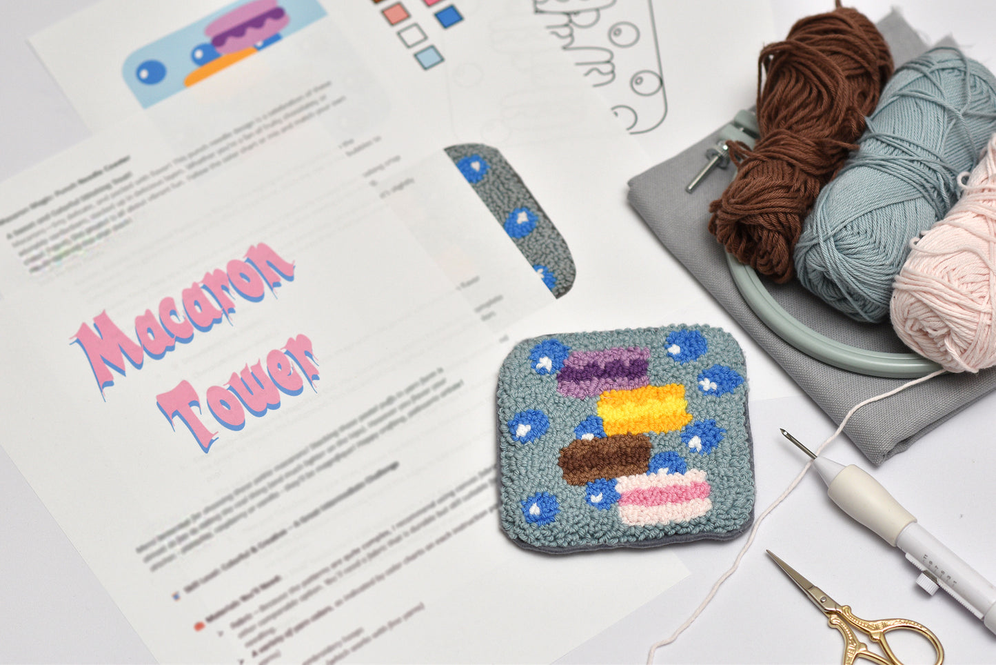 Embroidered square coaster with colorful macaron design on a table with yarn and a pattern sheet.