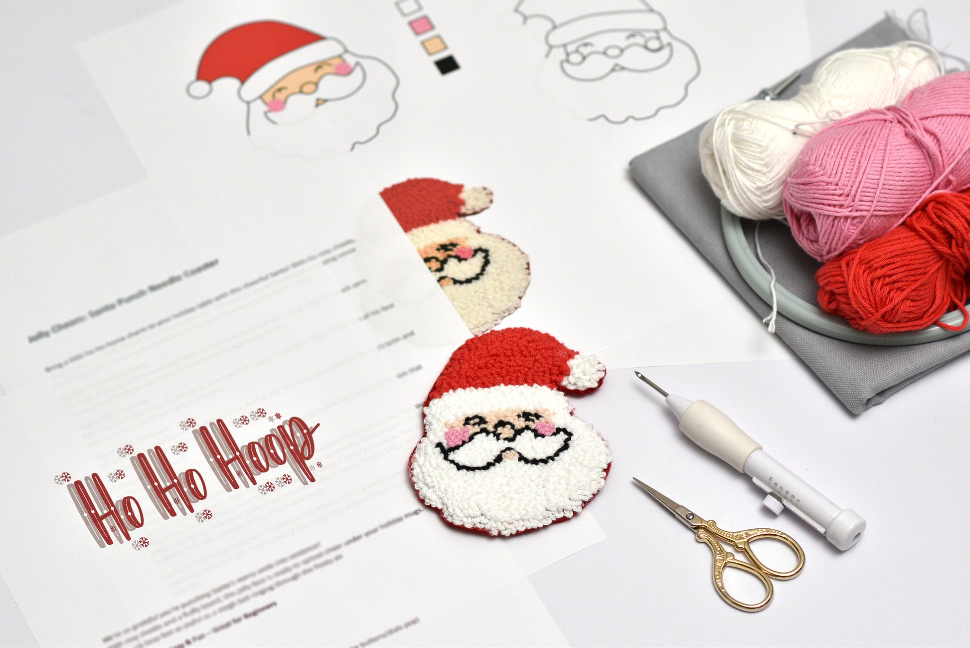 Punch needle materials with Santa Claus coaster design, yarn, scissors, and a punch needle hook on a white surface.