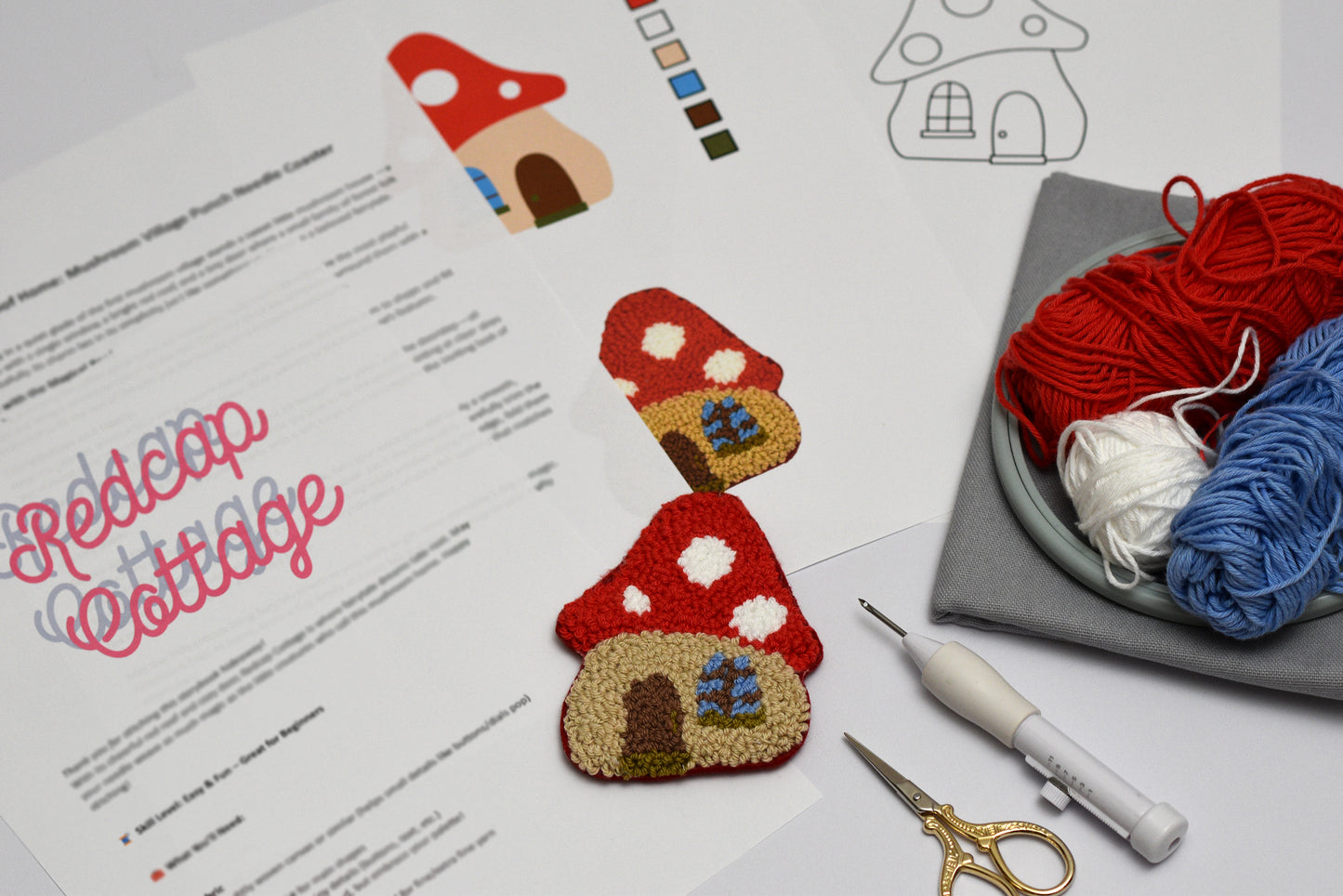 Embroidered  punch needle coaster with mushroom shaped house design on paper with yarn and sewing tools.