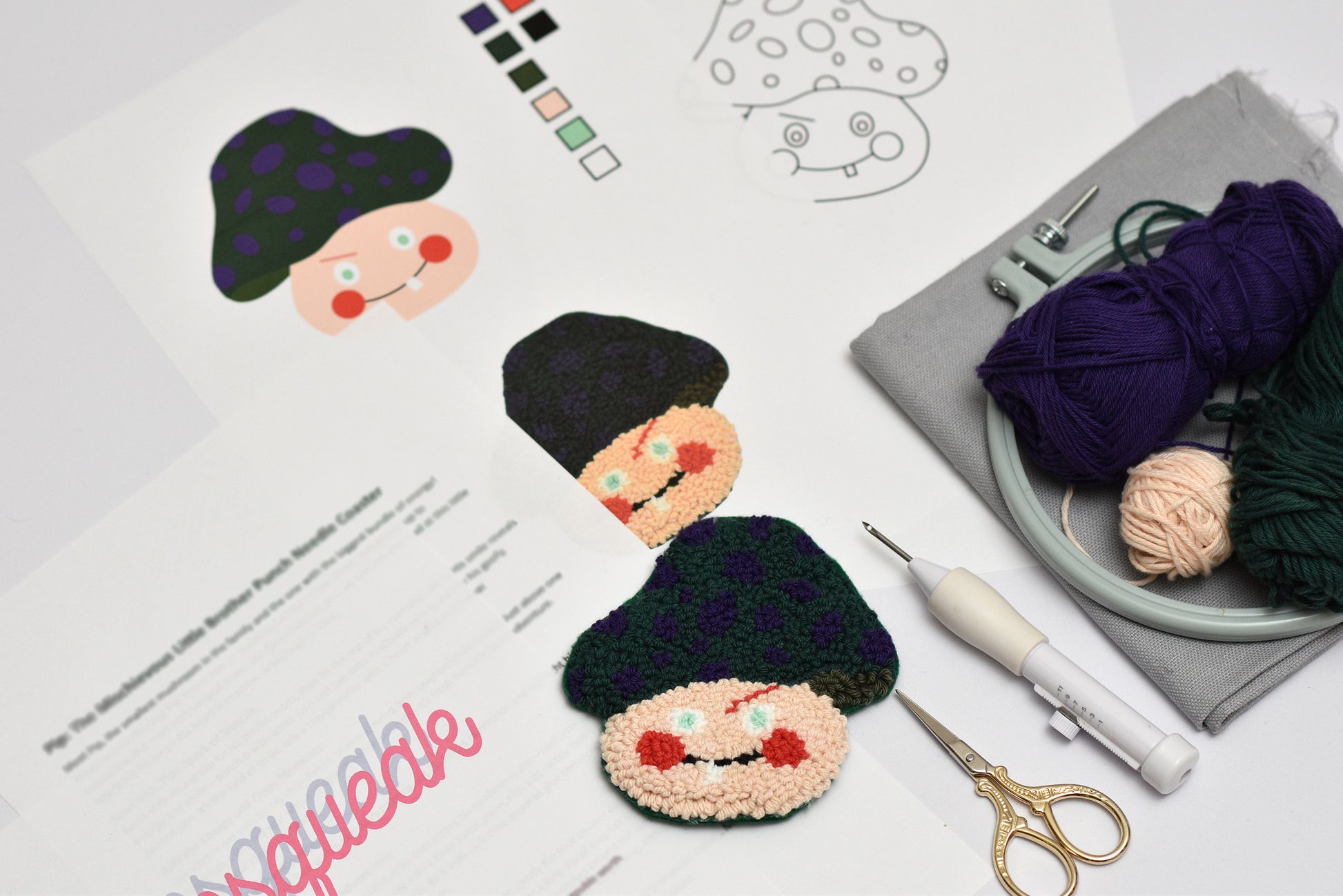 Embroidered coasters of mushroom cartoon characters with yarn, scissors, and a punch needle hook on a table.