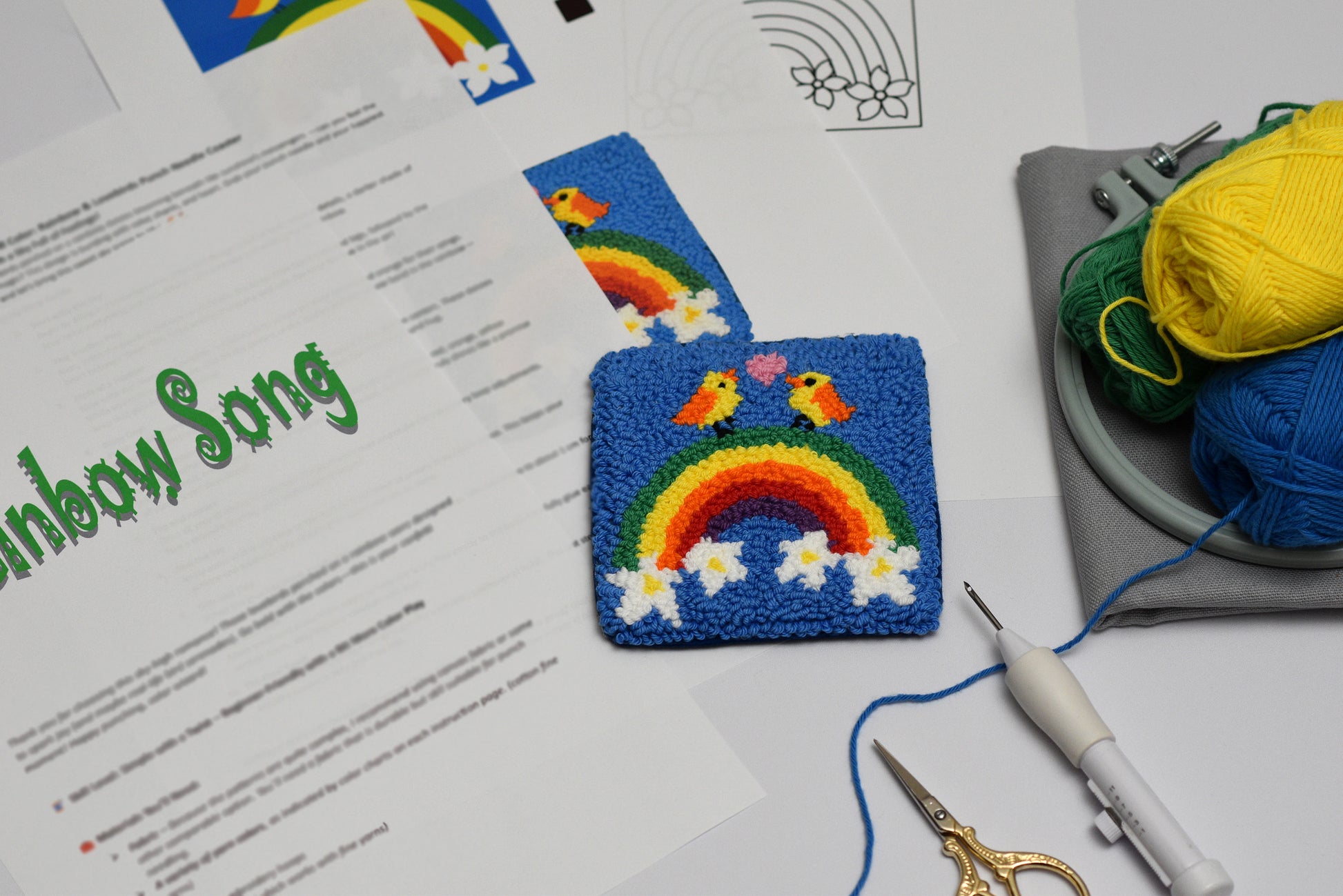 Handmade coaster with rainbow and bird design on a blue fabric square with crafting tools and materials.