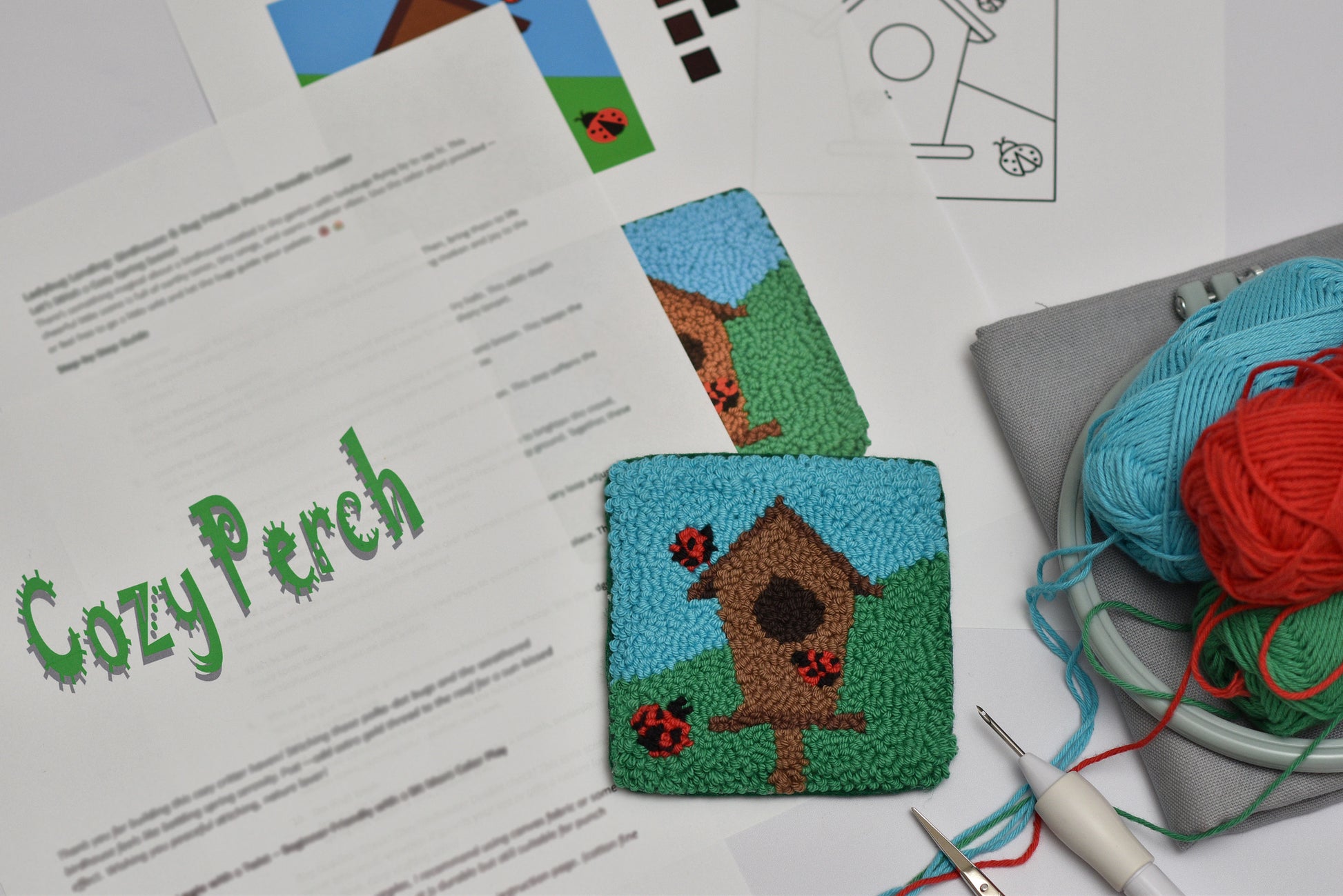 Embroidered mug rug with a birdhouse design on a piece of paper labeled 'Cozy Perch', surrounded by yarn and knitting tools.