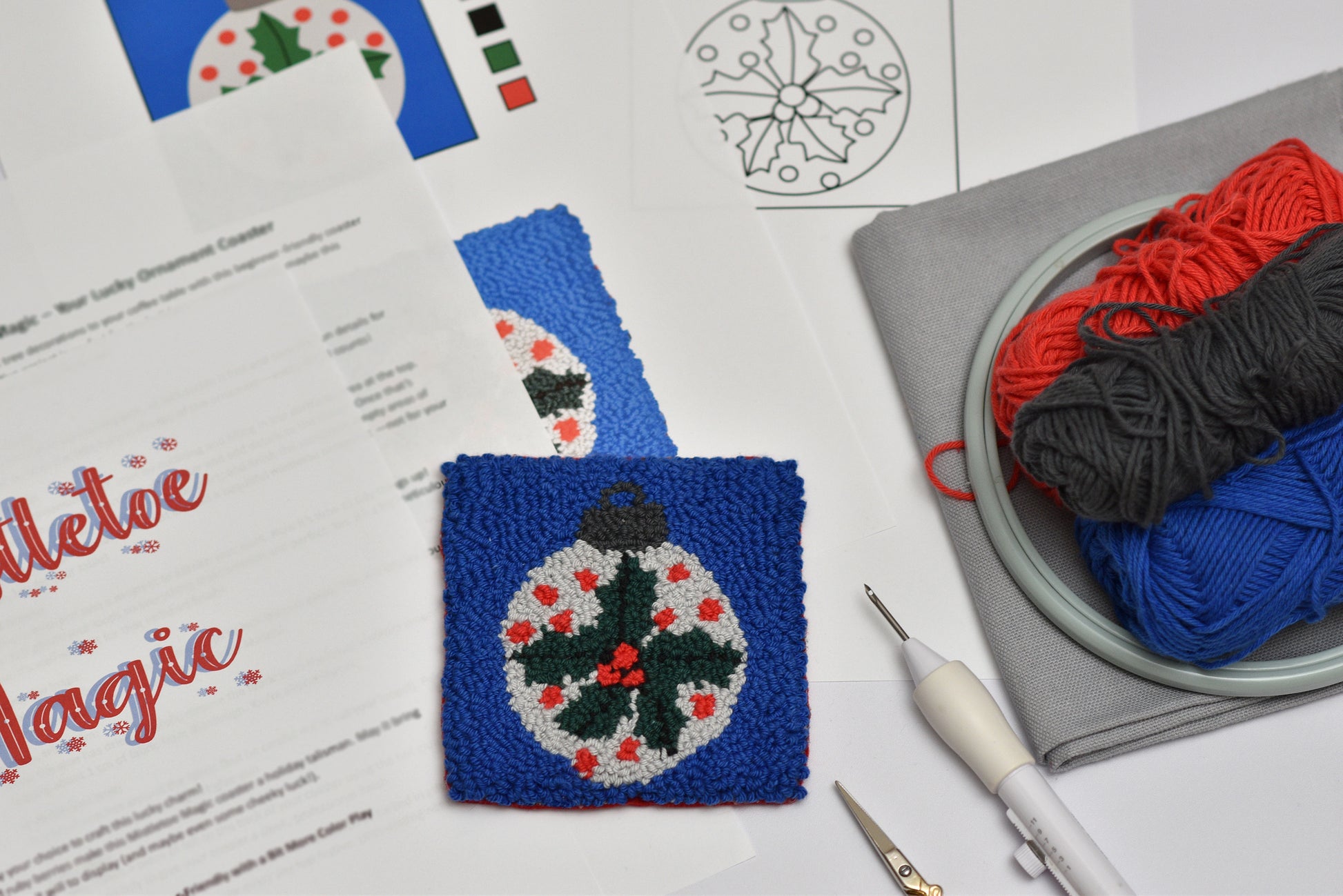 Punch needled coaster with mistletoe design on a table with yarn and patterns.