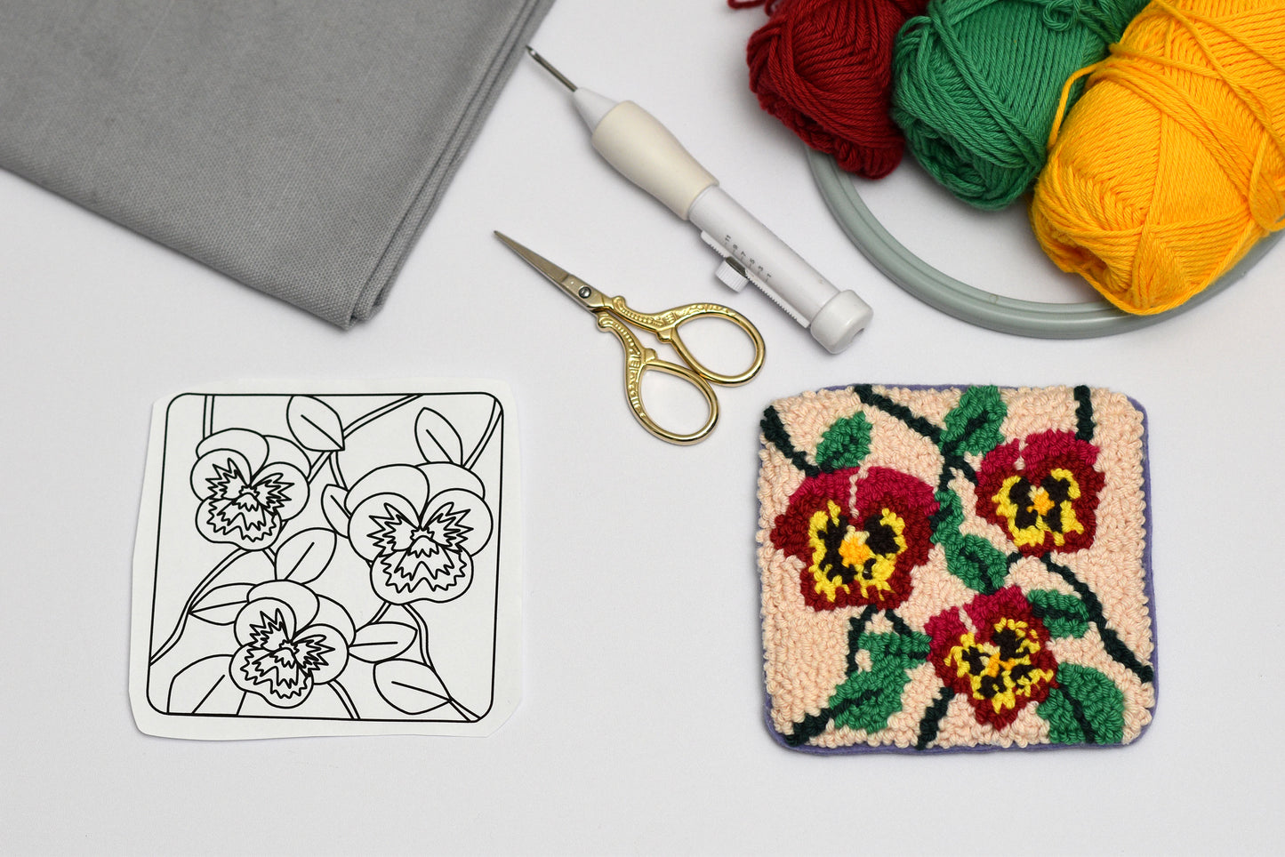 Embroidery hoop with floral design coaster, colorful yarn, fabric, and embroidery tools on a white surface.