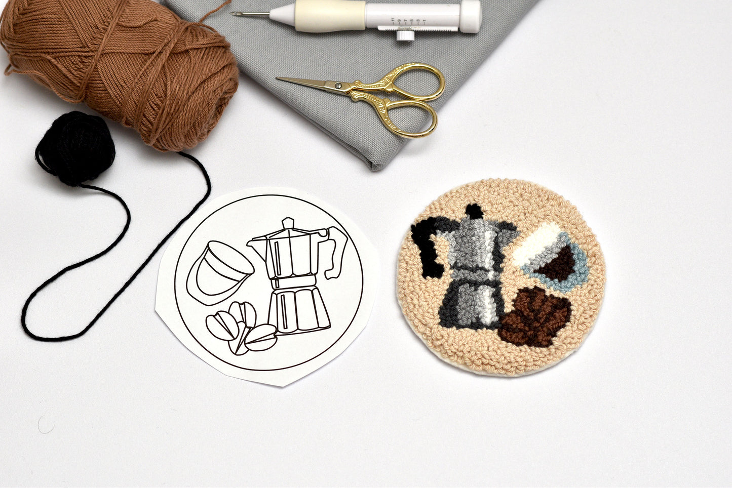 Embroidery hoop with a completed design of a coffee maker, surrounded by embroidery supplies on a white background.