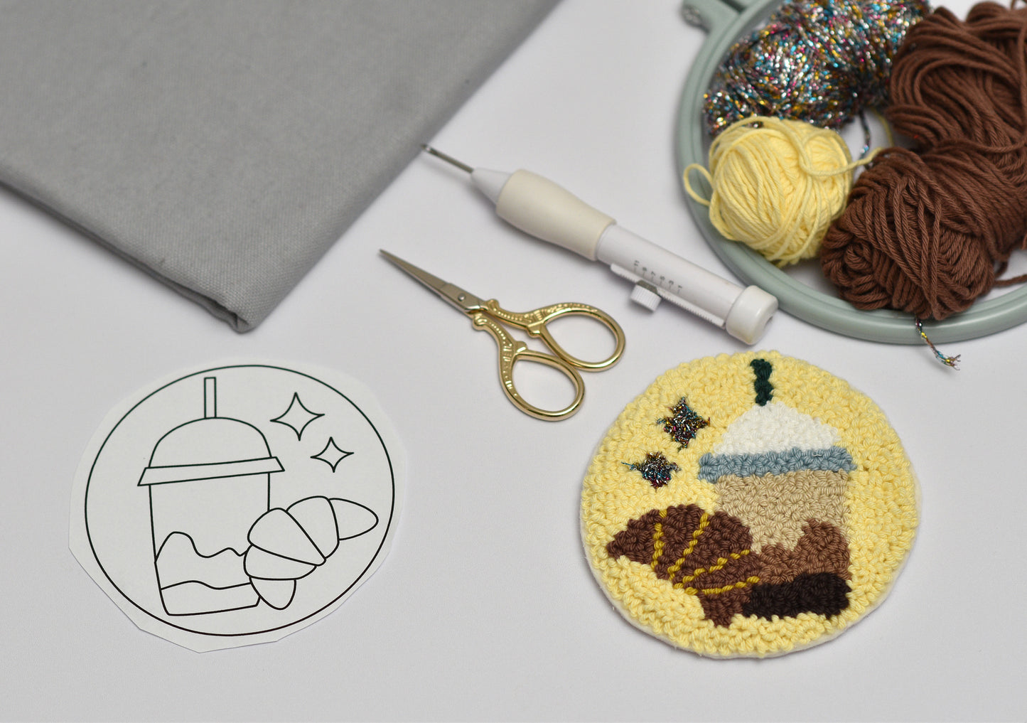 Embroidery hoop with yarn, scissors, and a completed embroidery coaster with coffee and Croissant design on a white surface.