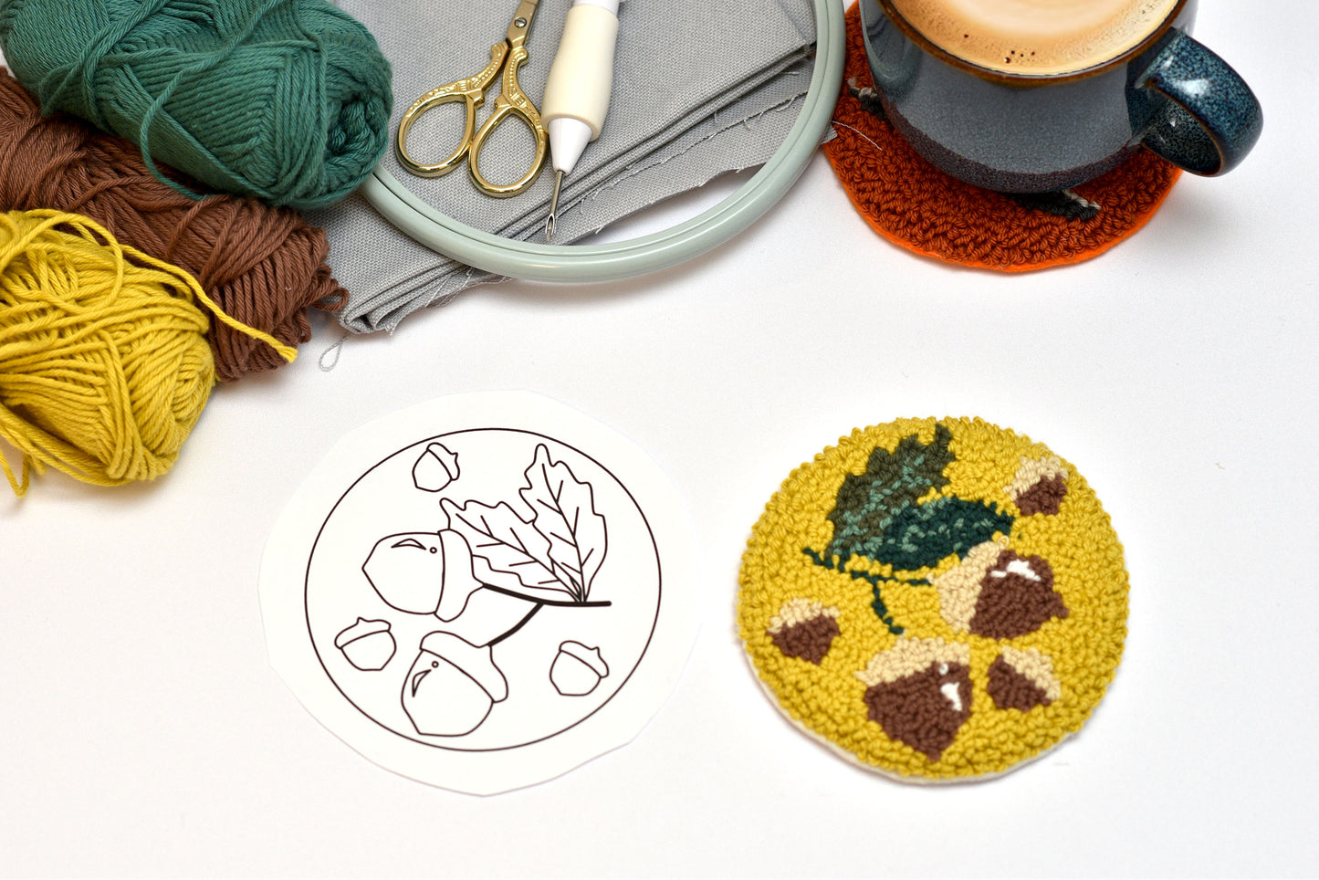 Coaster with leaf and hazelnut design, yarn, scissors, and a cup on a white surface