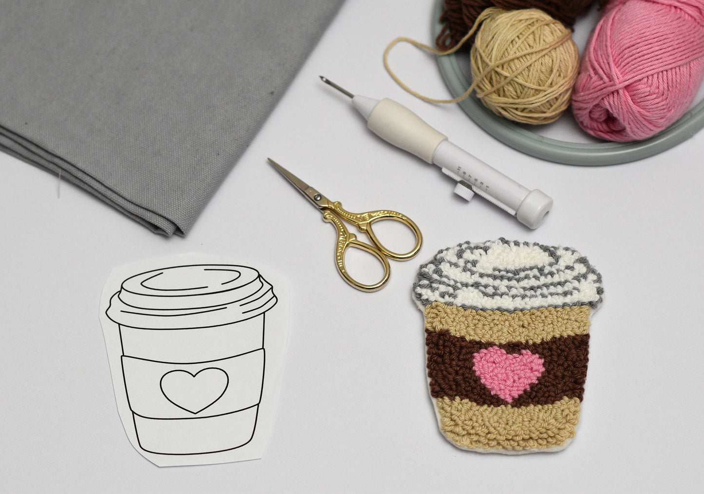 Punch needled coffee cup coaster with pink heart next to a line drawing of a coffee cup on a table with yarn and tools.