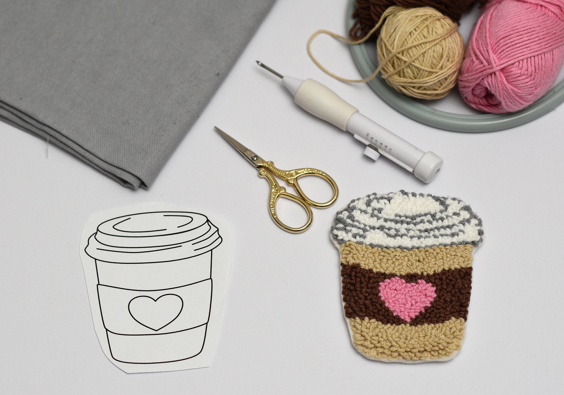 Punch needled coffee cup coaster with pink heart next to a line drawing of a coffee cup on a table with yarn and tools.