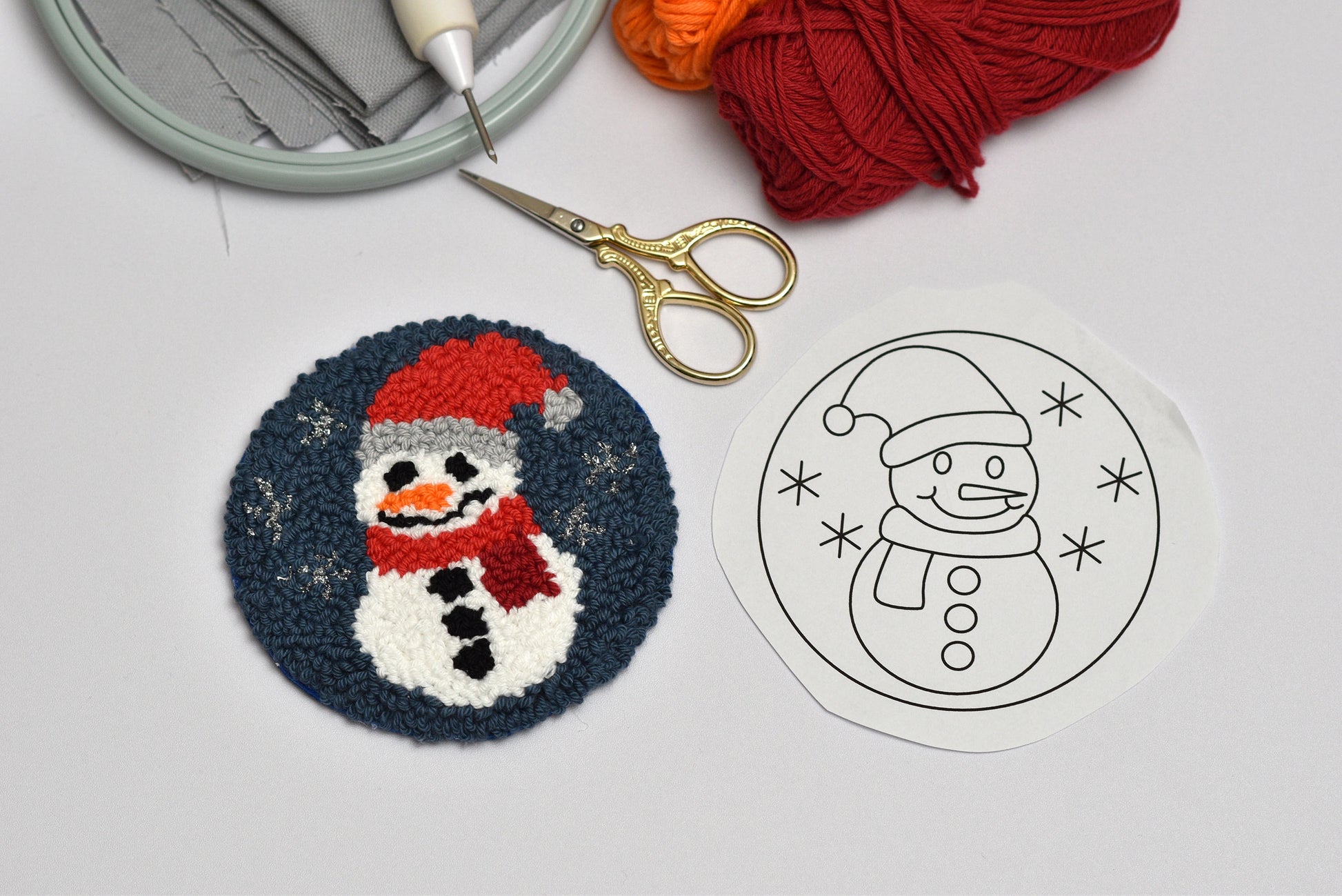 Embroidery hoop with yarn, scissors, and a snowman-themed punch needle design on a light gray background.