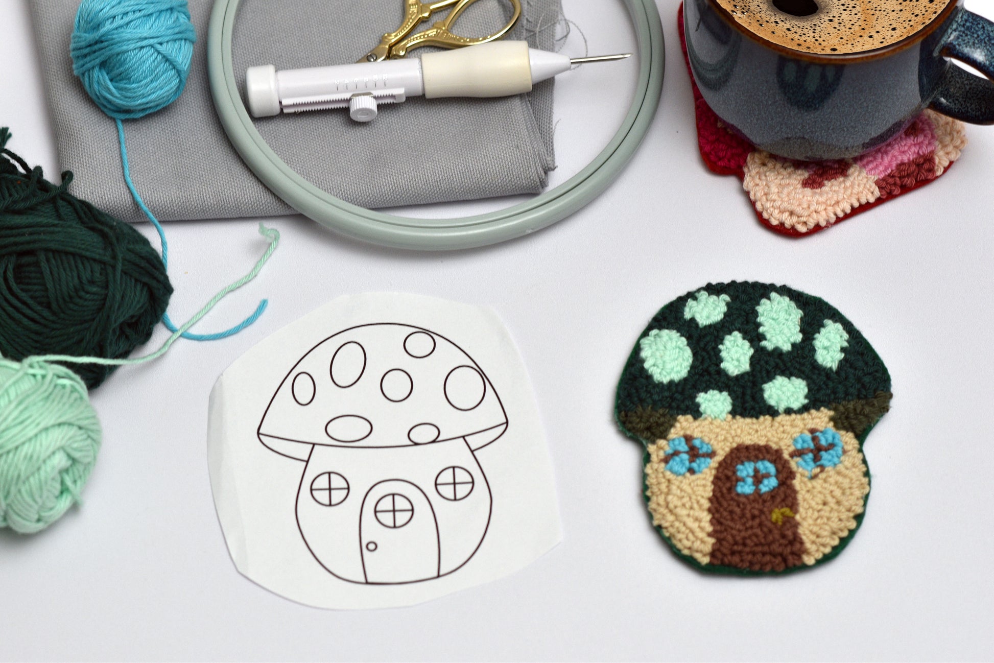 Embroidery hoop with yarn, scissors, and a completed mushroom house coaster and its design on a white surface.