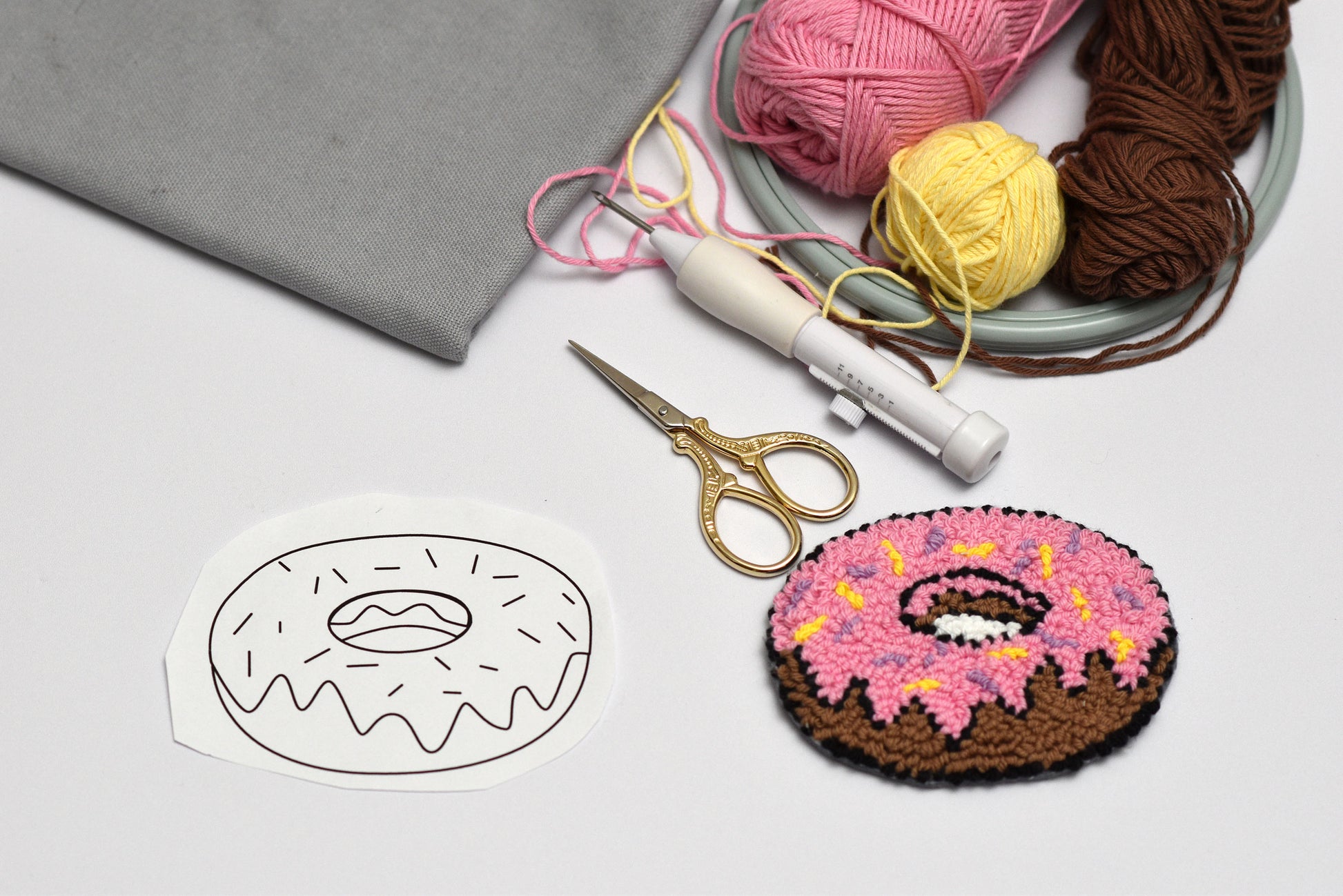 Punch needle donut with pink icing and brown base, next to yarn balls and a template on a light gray background.