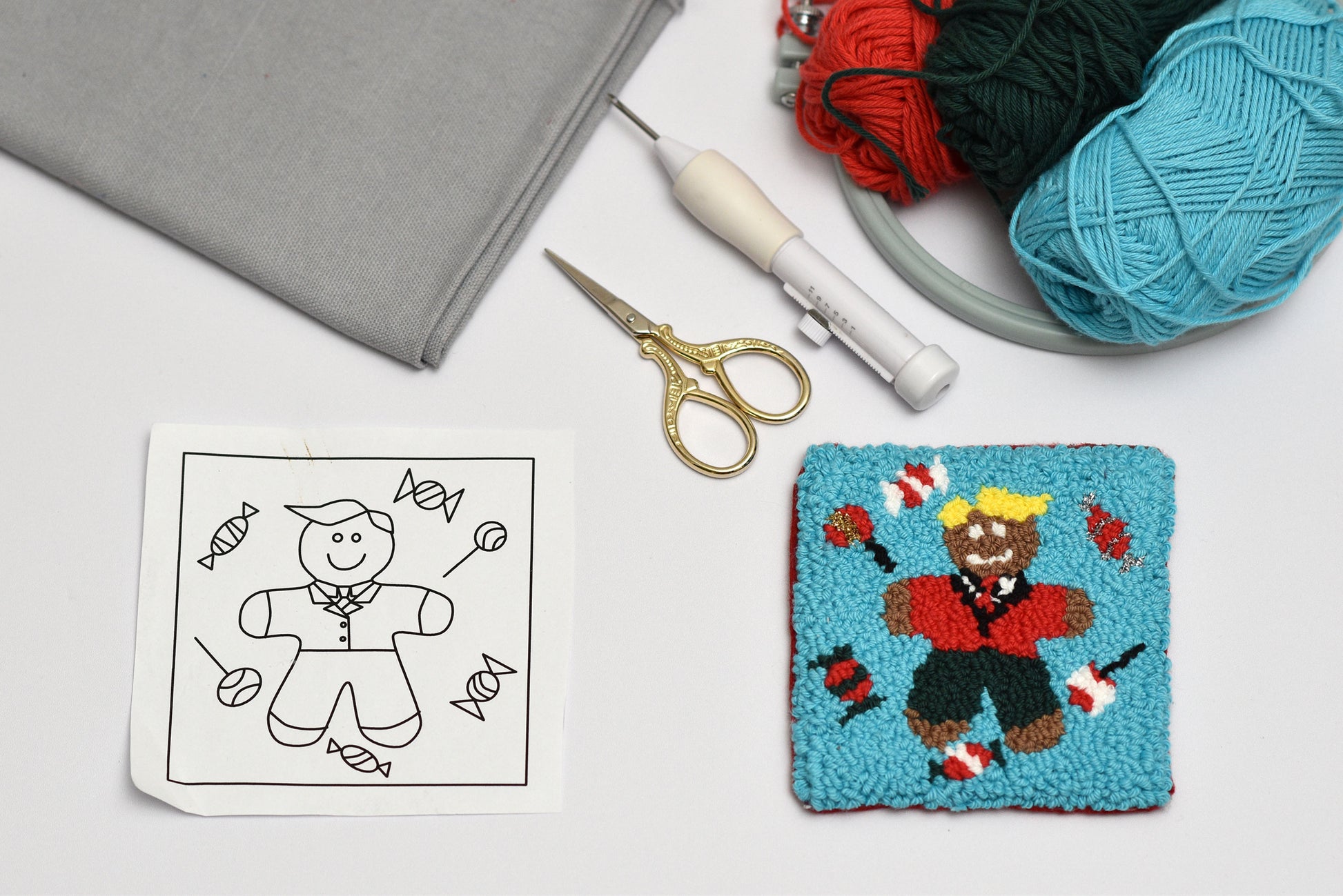 Embroidery coaster project with a completed patch of a gingerbread man holding a candy, a pattern sheet, scissors, and yarn on a white surface.