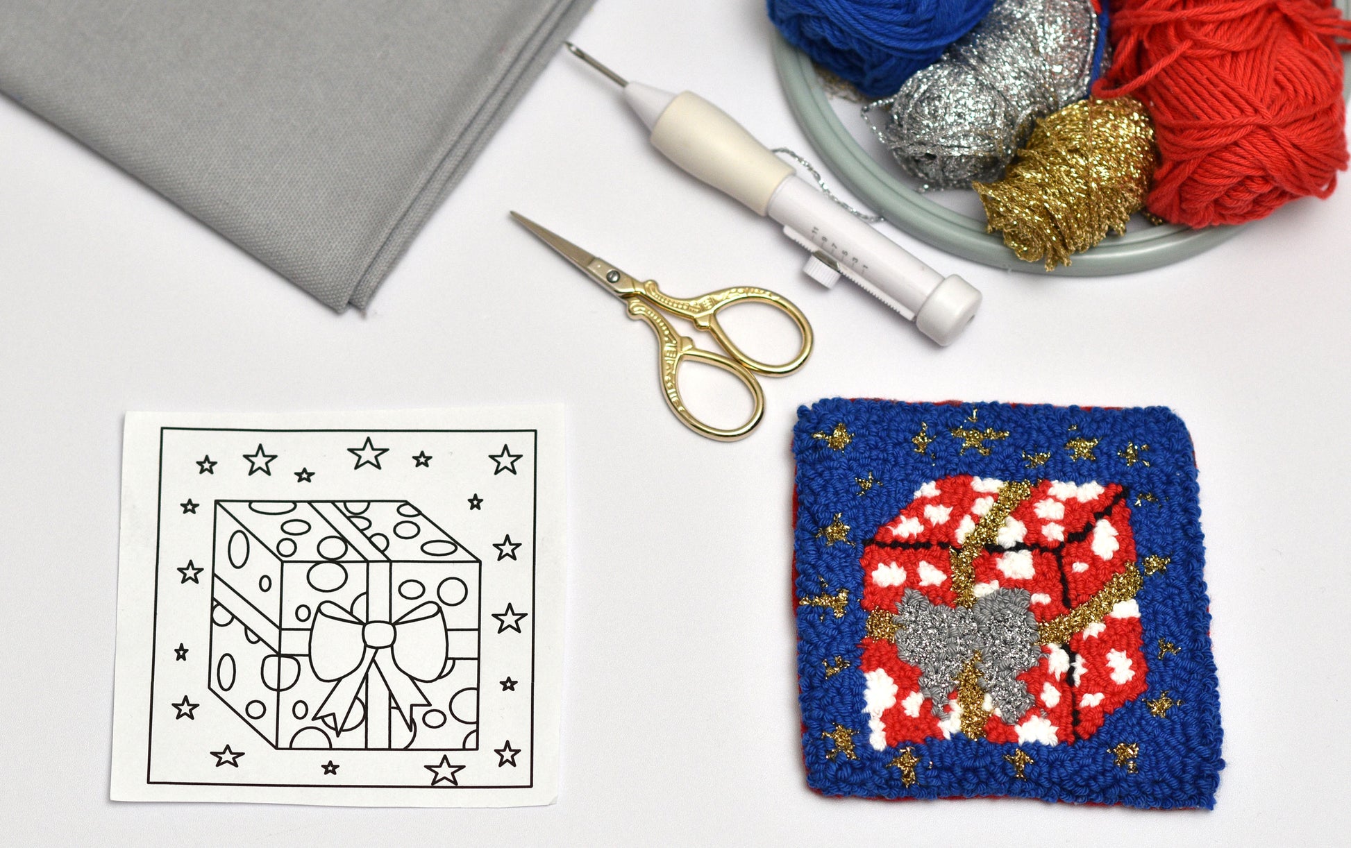 Punch needled square coaster with gift package design next to a pattern sheet, scissors, and yarn on a white surface.