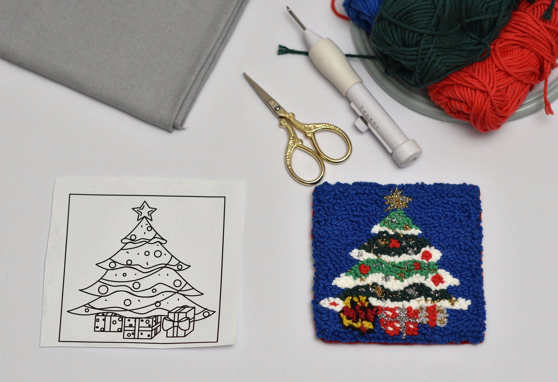 Embroidery coaster project with a Christmas tree design, yarn, scissors, and fabric on a table.