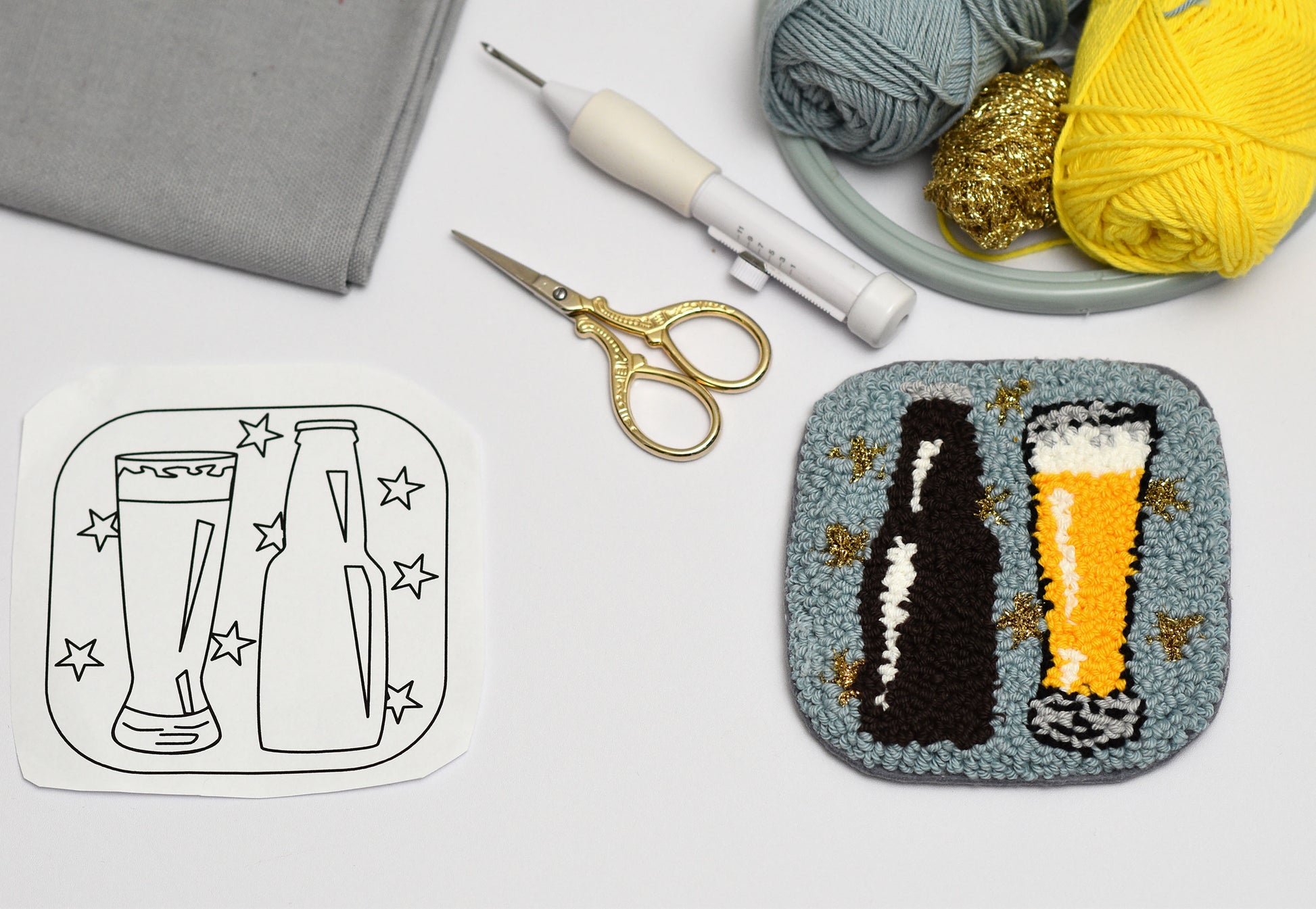 Embroidery coaster project with beer-themed design, scissors, and yarn on a white surface