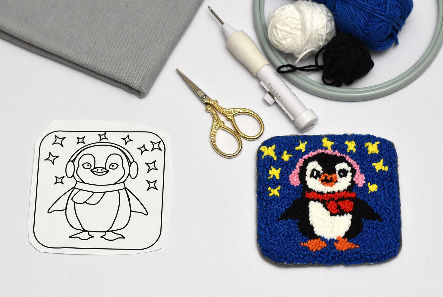 Punch needled penguin coaster next to a black and white outline of a penguin, with yarn and scissors on a white surface.