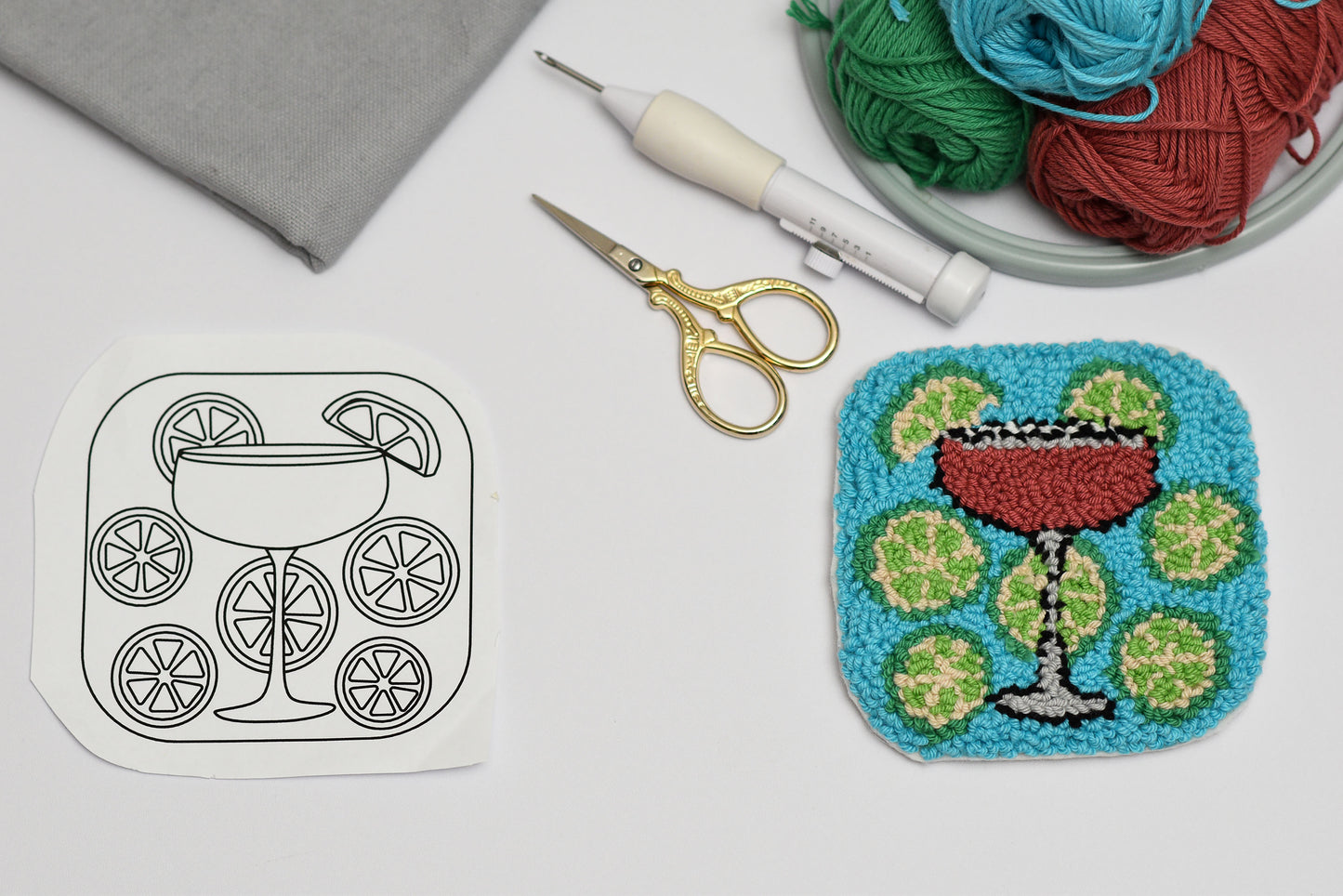 Embroidery hoop with a completed punch needled design of a cocktail cosmopolitan, surrounded by embroidery supplies on a white surface.