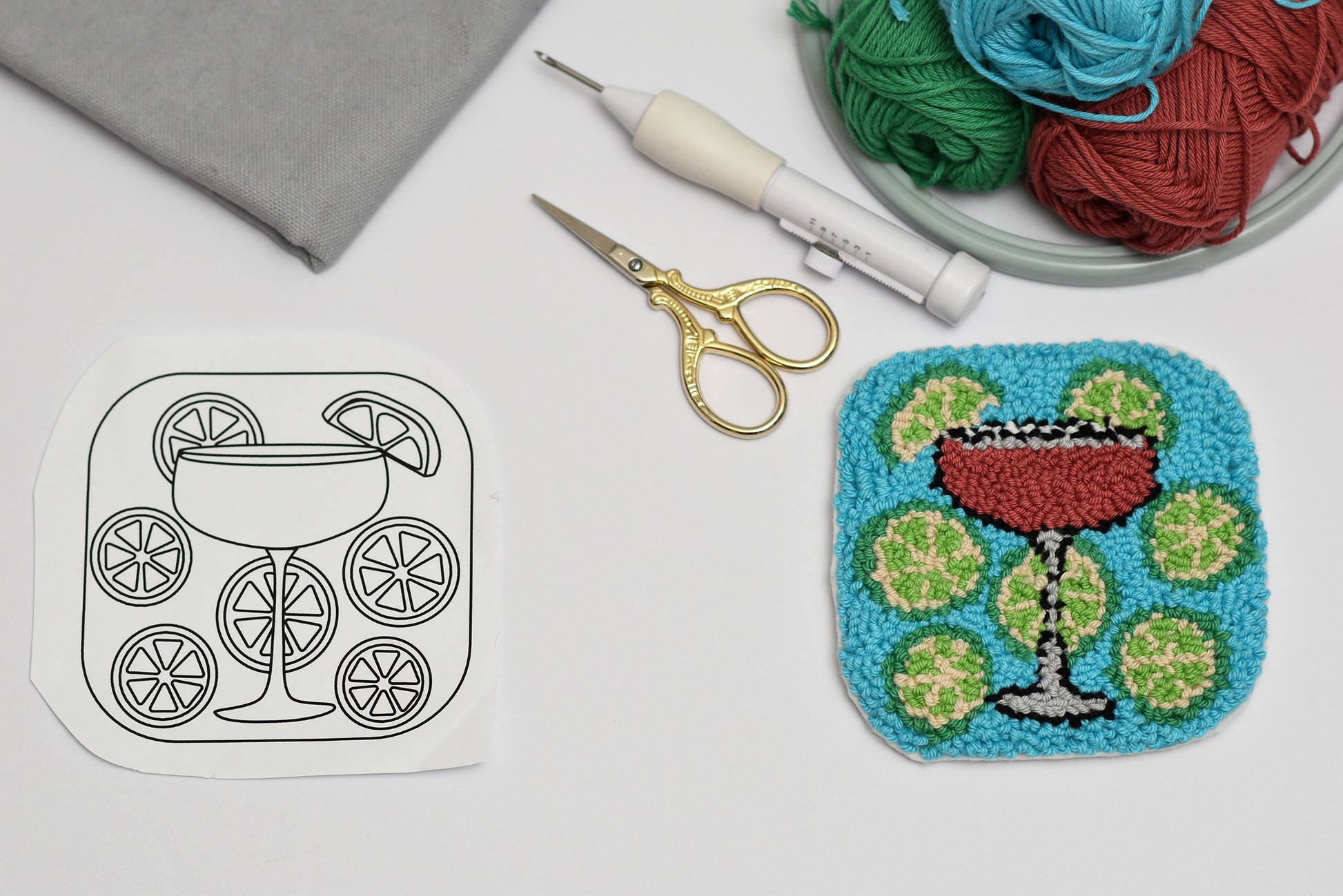 Embroidery hoop with a completed punch needled design of a cocktail cosmopolitan, surrounded by embroidery supplies on a white surface.