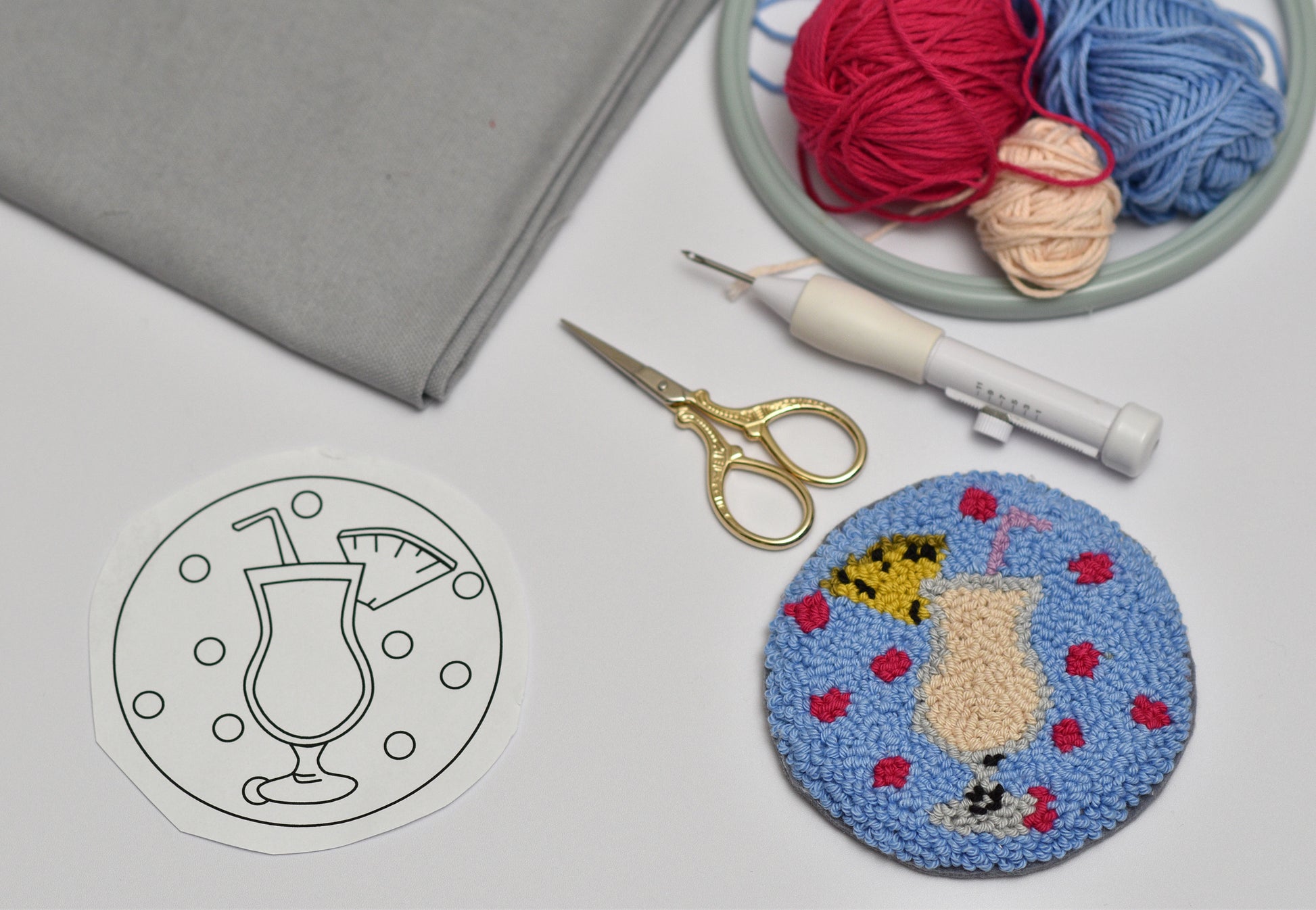 Embroidered coaster with cocktail design next to a coloring page, scissors, and yarn on a gray surface.
