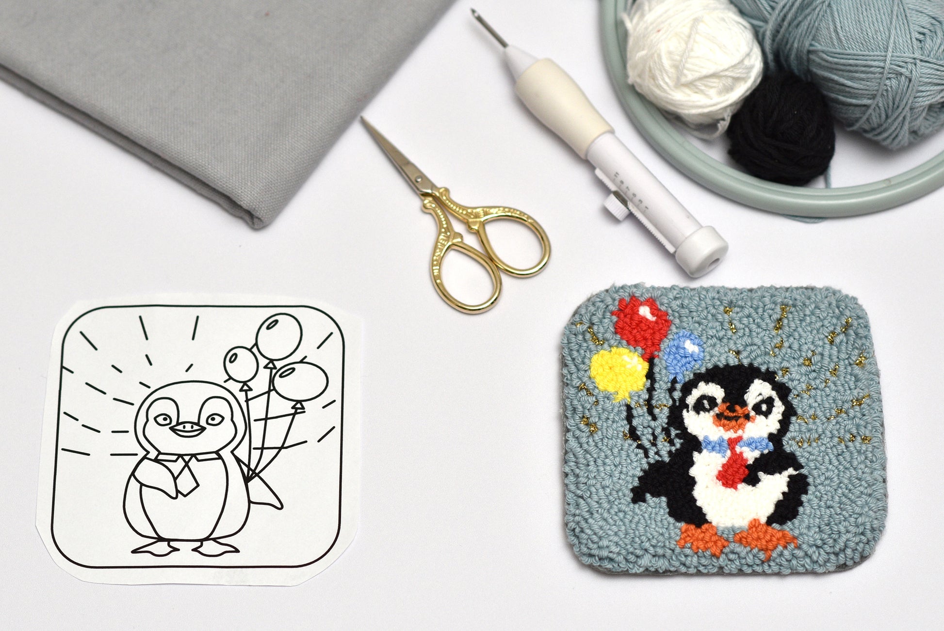 Punch needled coaster with design of a penguin with balloons next to a black and white outline on a white surface with yarn and scissors.