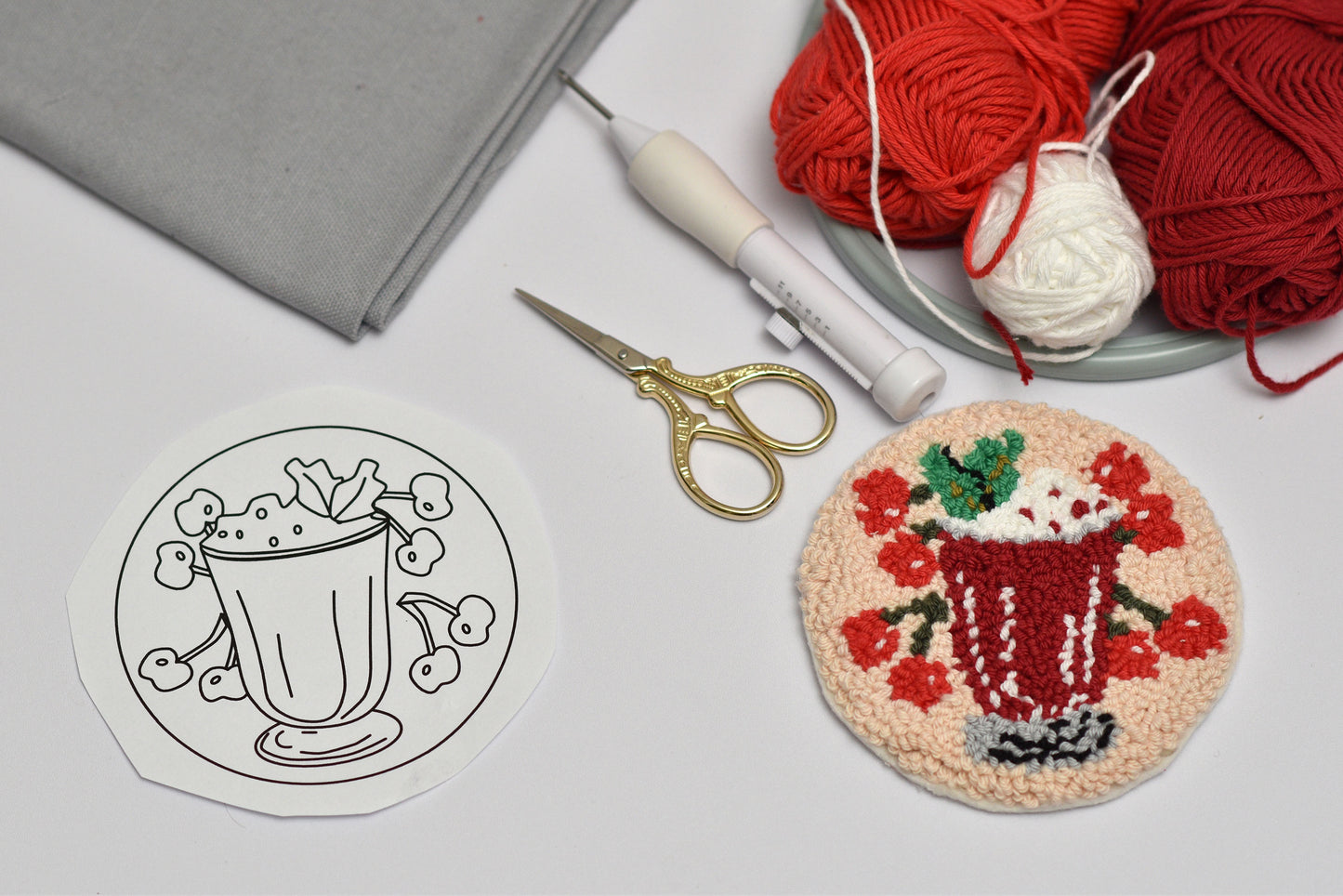 Embroidery hoop with a completed cherry cocktail coaster design, yarn, scissors, and gray fabric on a light surface