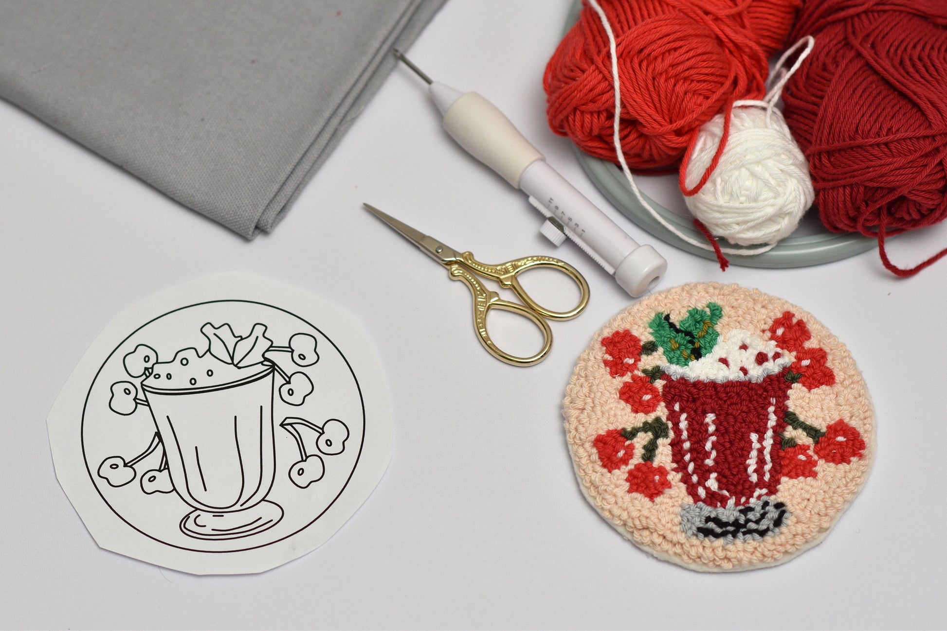 Embroidery hoop with a completed cherry cocktail coaster design, yarn, scissors, and gray fabric on a light surface