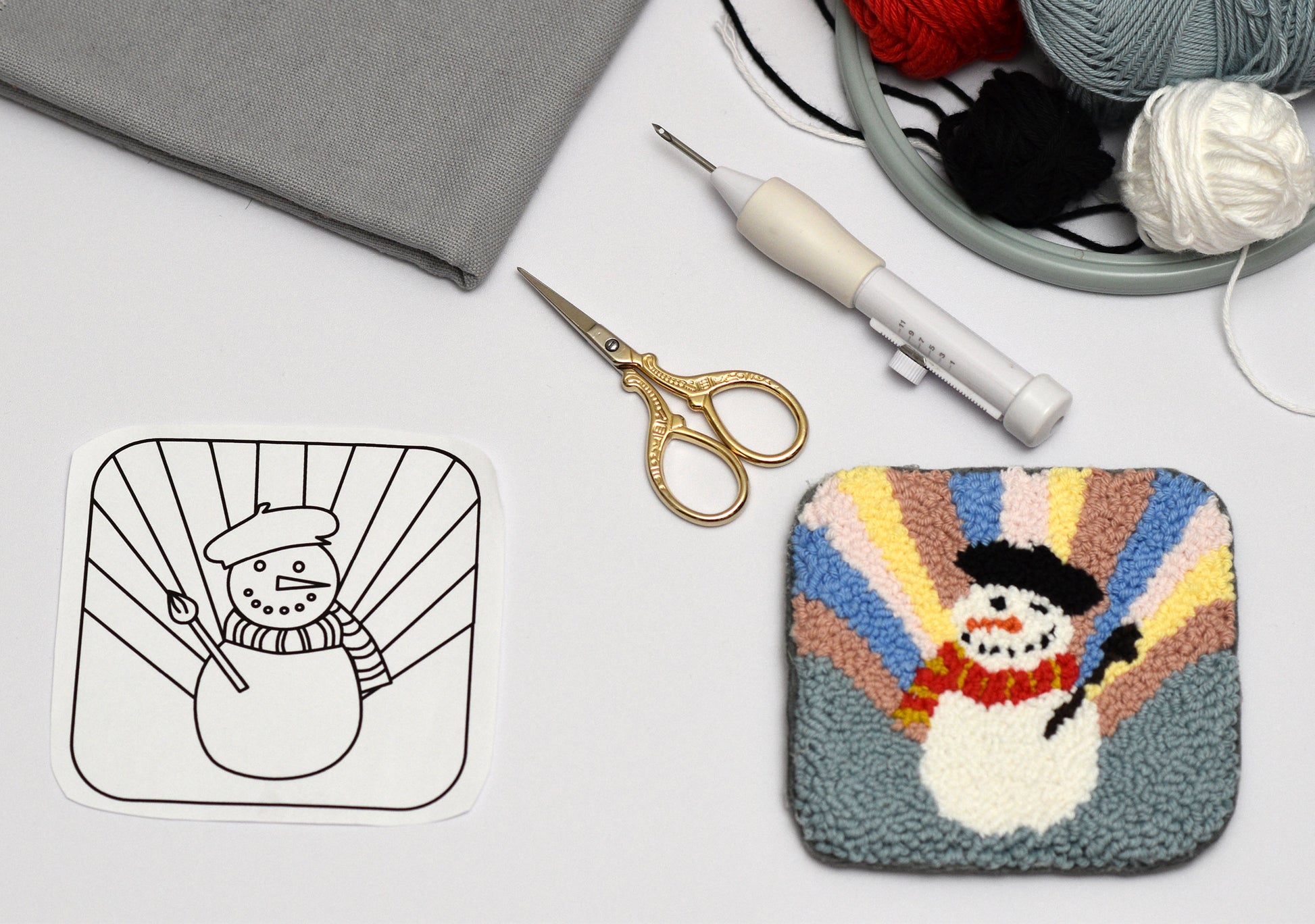 Colorful punch needle coaster with snowman design next to a black and white outline, scissors, and yarn on a light surface.