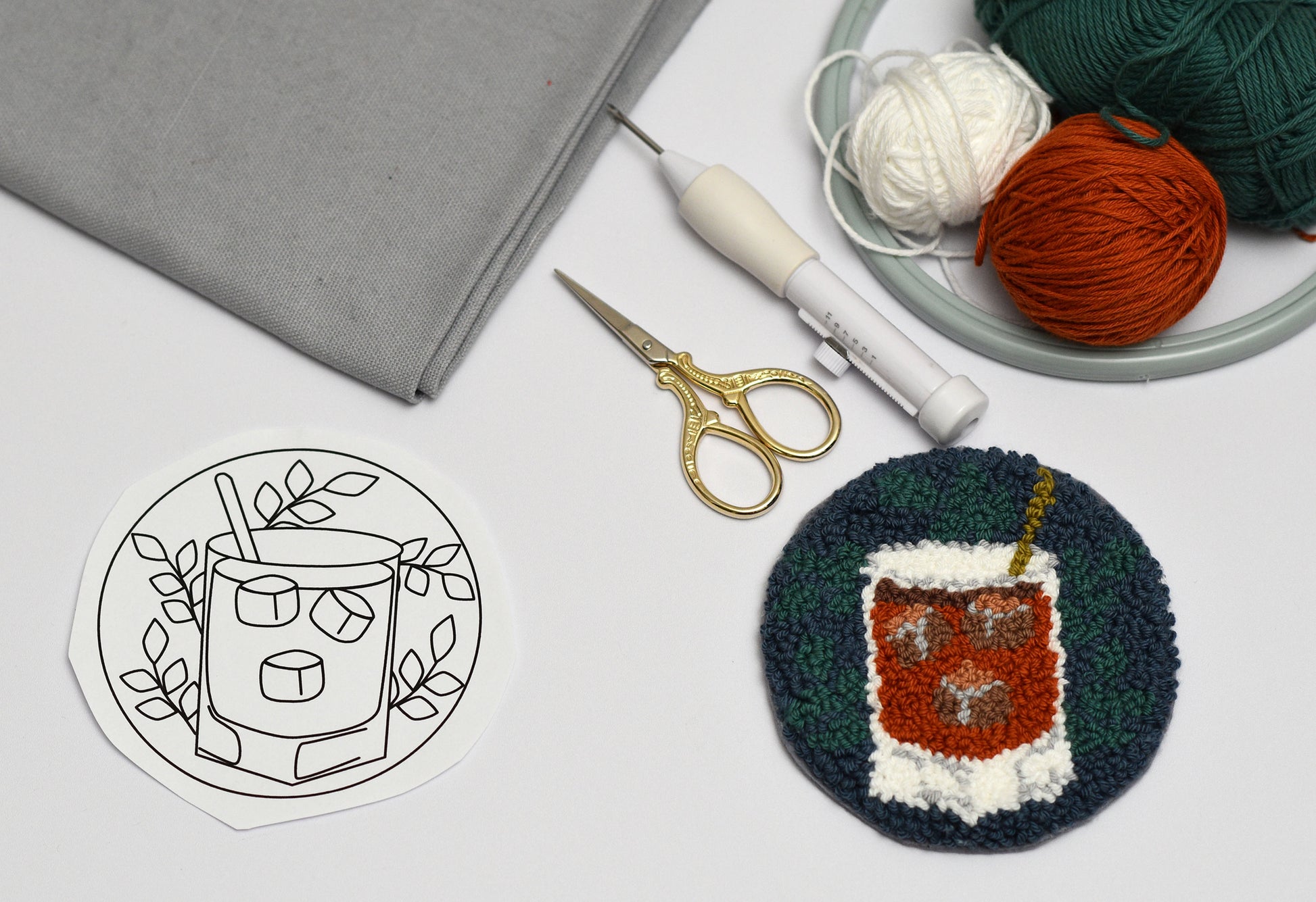 Embroidery hoop with a completed design of a Whiskey Coaster, next to a template, scissors, and yarn on a white surface.