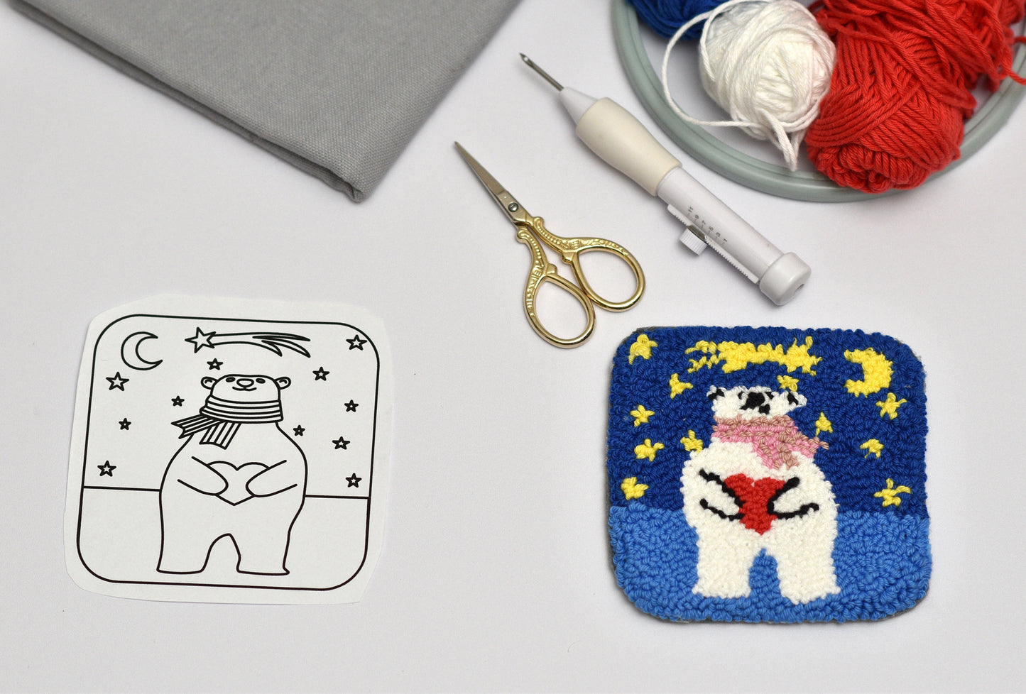 Embroidery coaster project with a cartoon bear design on fabric, surrounded by embroidery tools and yarn.
