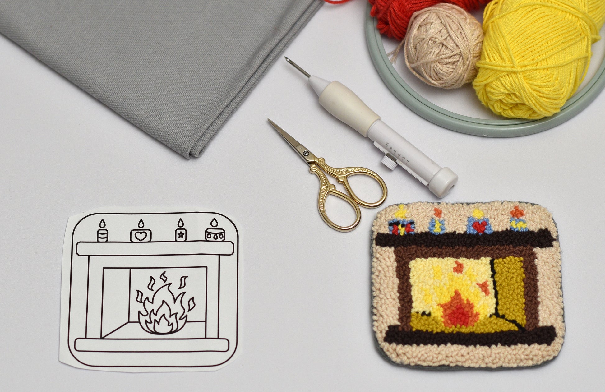 Punch needled coaster pattern with fireplace design next to yarn, scissors, and a gray fabric background